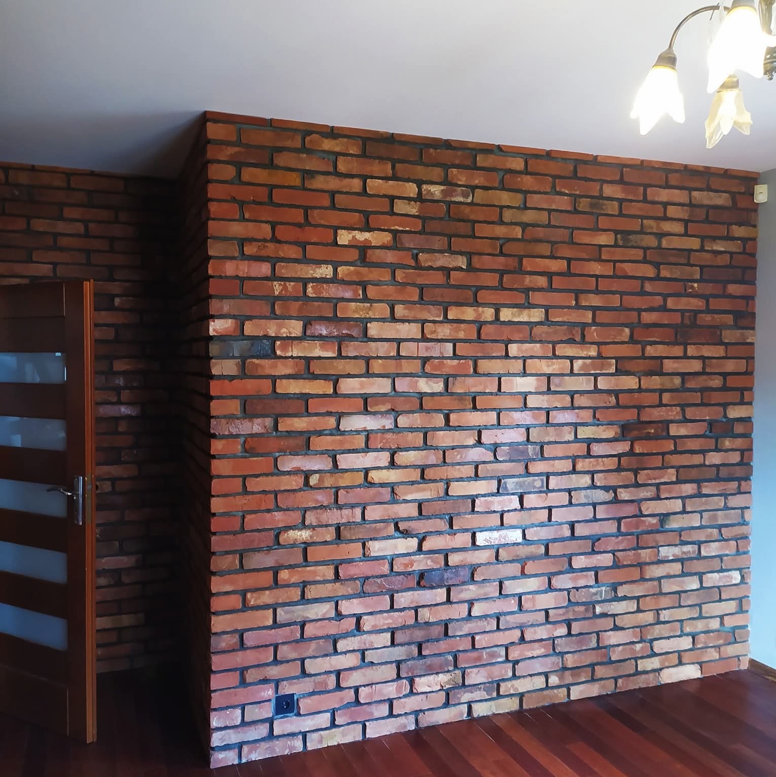 Ściana z cegły klinkierowej w salonie z drewnianą podłogą i częściowo otwartymi drzwiami. Widoczna lampa sufitowa i gładka ściana boczna.