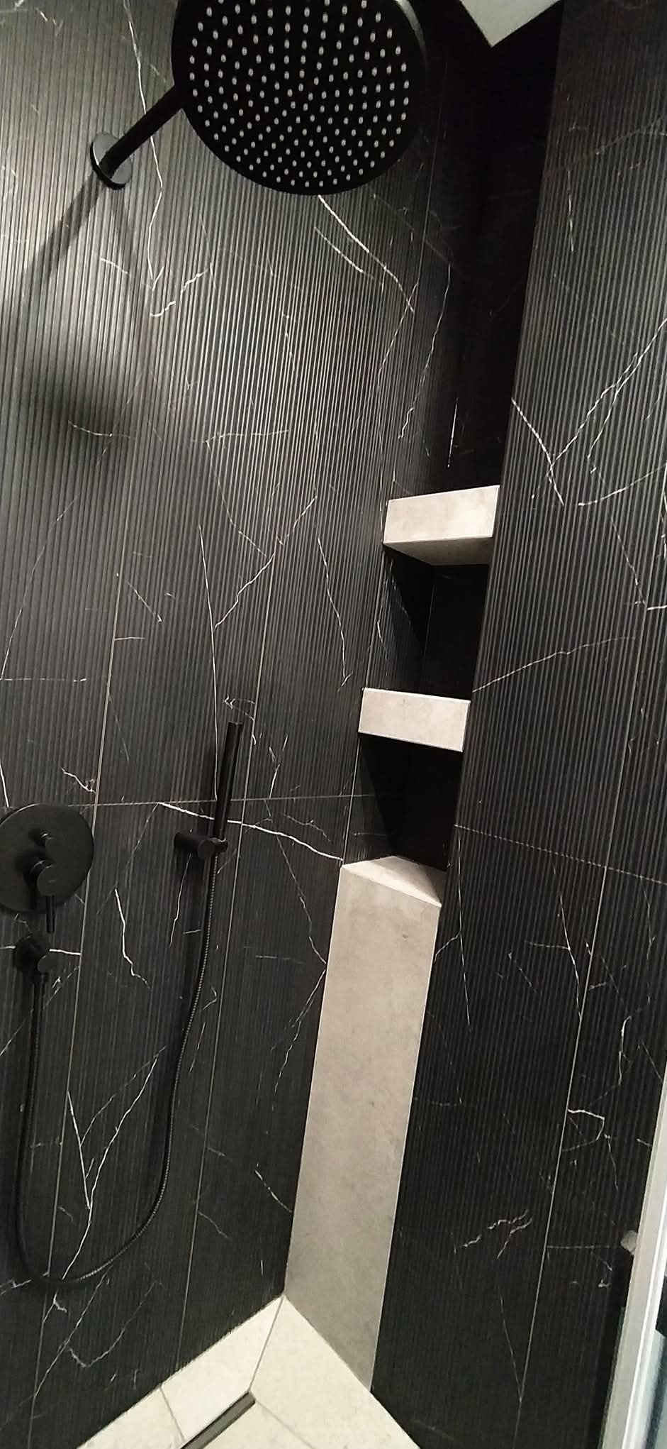 Nowoczesna kabina prysznicowa z czarnymi płytkami imitującymi marmur i wbudowanymi półkami w kolorze szarości. Minimalistyczna armatura w czarnym macie.