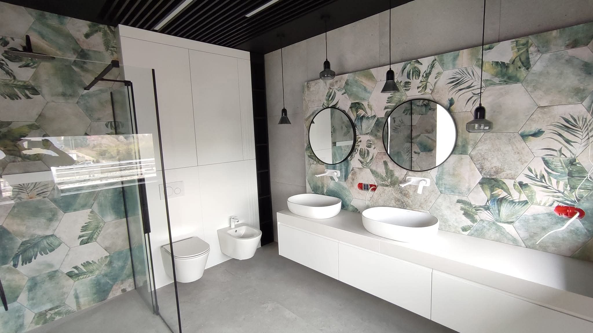 Nowoczesna łazienka z heksagonalnymi płytkami w tropikalne wzory, okrągłe lustra, białe umywalki nablatowe i kabina prysznicowa ze szkła.