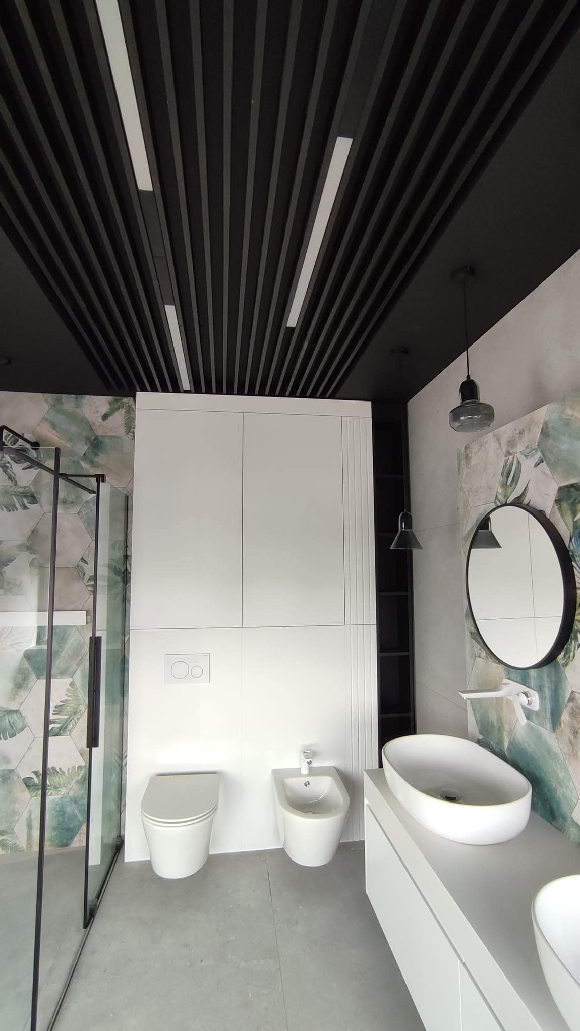 Nowoczesna łazienka z kabiną prysznicową, toaletą, bidetem i umywalką nablatową. Czarny sufit lamelowy z oświetleniem. Ściany z motywem roślinnym.