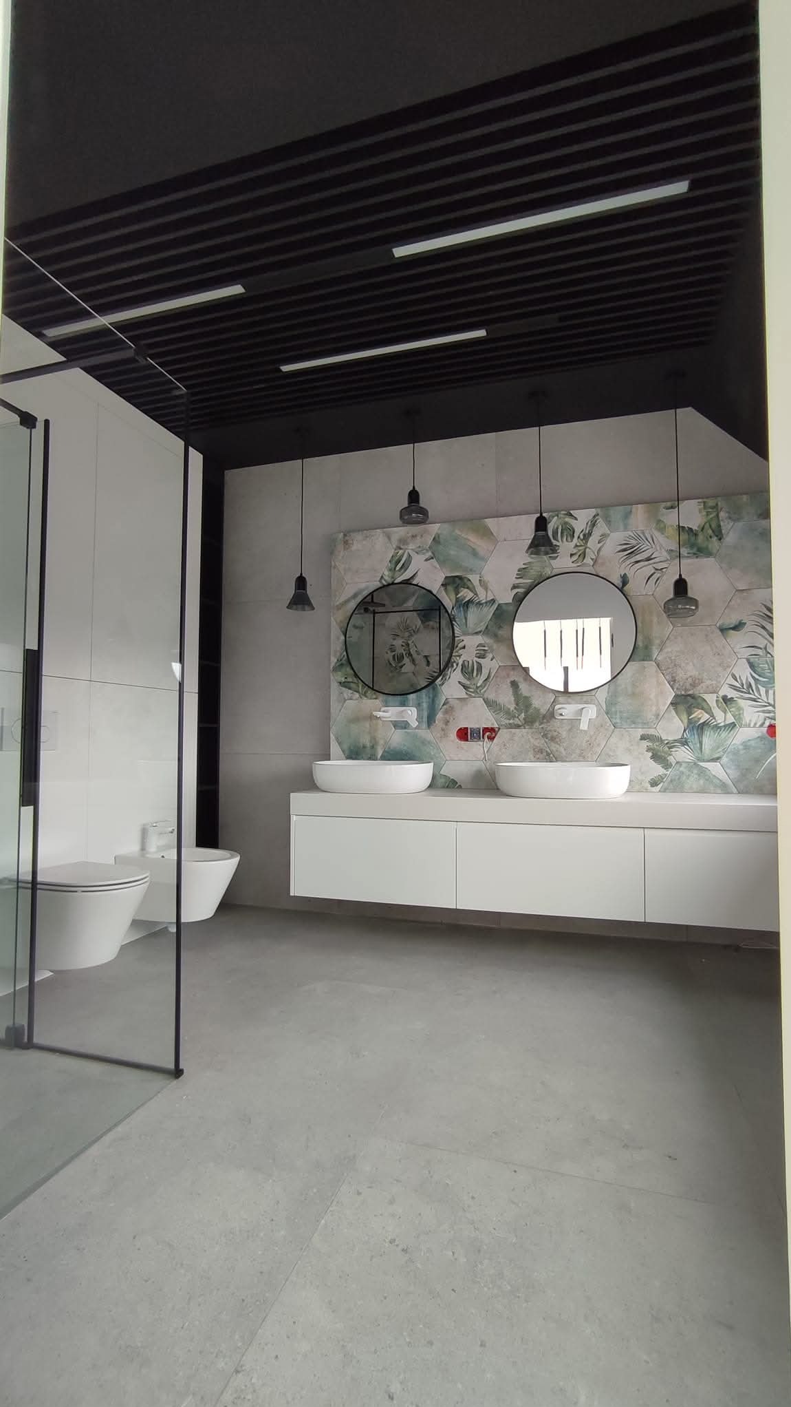 Nowoczesna łazienka z podwójną umywalką, okrągłymi lustrami i heksagonalnymi płytkami w roślinny wzór. Czarna, ażurowa konstrukcja sufitu z oświetleniem.