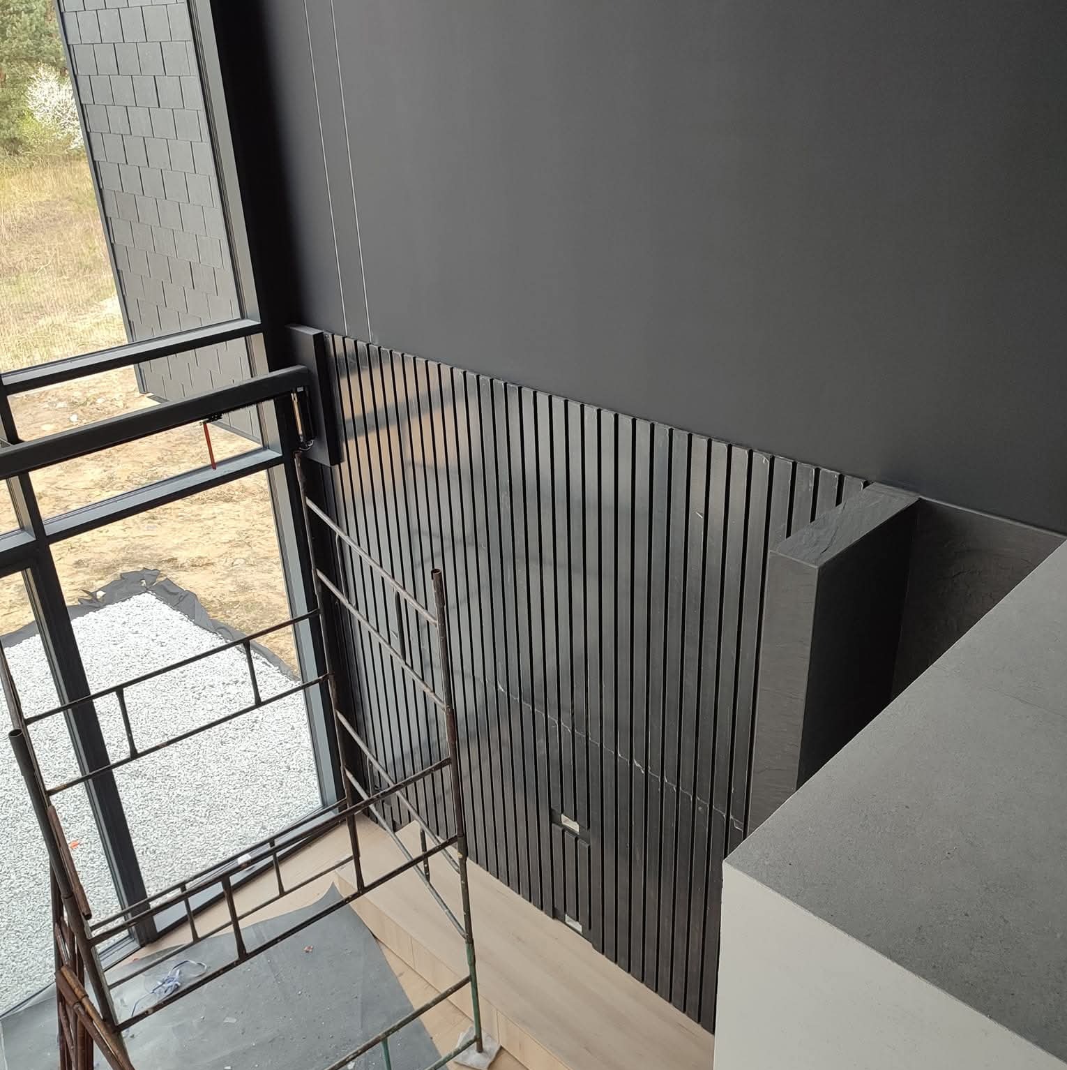 Wysoki pokój z szarą ścianą i panelami dekoracyjnymi. Widoczne okno z widokiem na zewnątrz i rusztowanie w rogu. Minimalistyczny design wnętrza w odcieniach szarości.