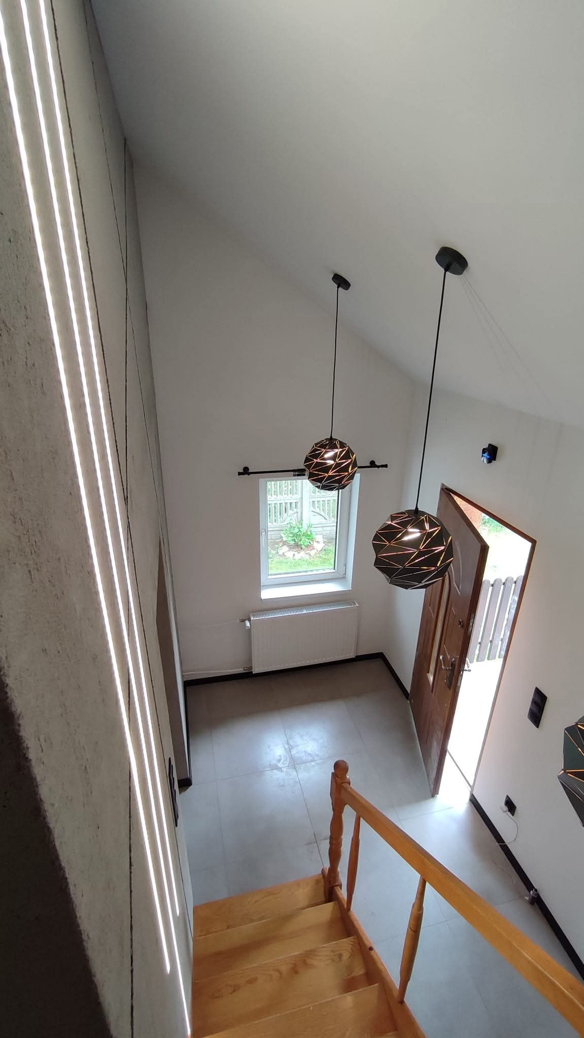 Nowoczesne wnętrze domu z drewnianymi schodami, geometrycznymi lampami wiszącymi i oknem z widokiem na ogród, uchwycone z góry.