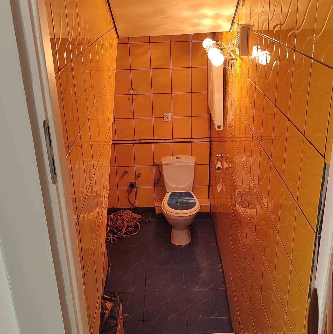 Pomarańczowa łazienka z niebieską podłogą, widok zza progu. Widoczna toaleta z deską z motywem morskim, grzejnik i kable wystające ze ściany.