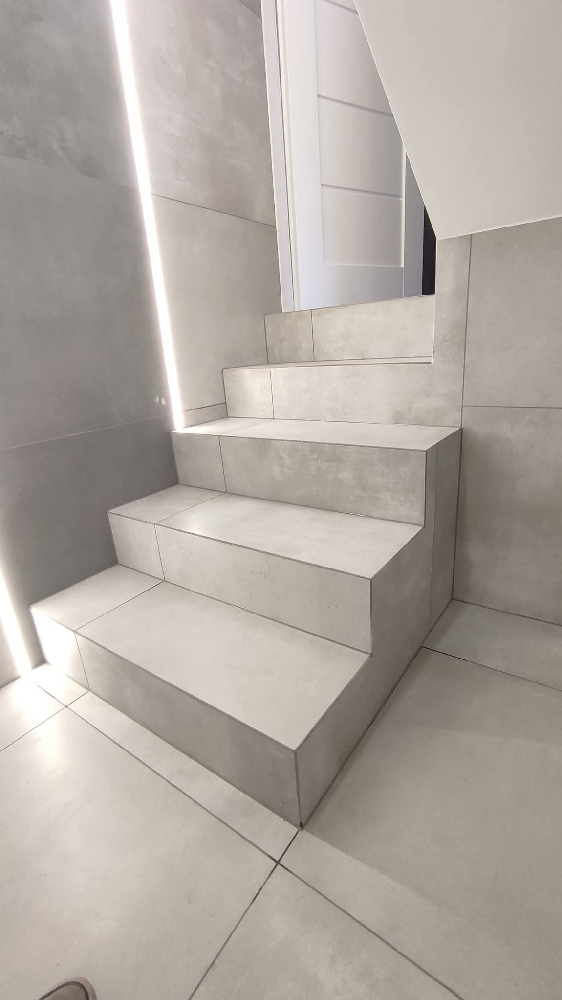 Schody wyłożone szarymi płytami ceramicznymi, z podświetleniem LED wzdłuż ściany. Minimalistyczny design, nowoczesne wykończenie wnętrza. Widok z dołu.