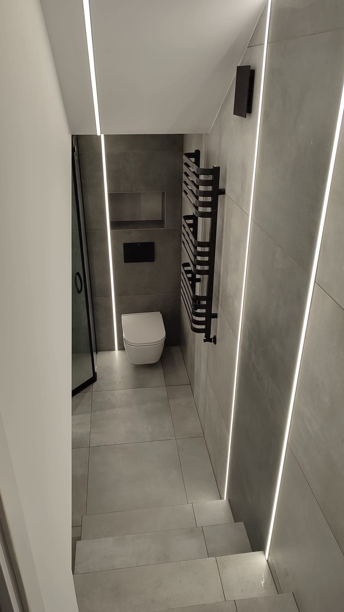 Nowoczesna łazienka w szarościach z podświetleniem LED. Toaleta wisząca, grzejnik drabinkowy i schody wykończone płytkami. Minimalistyczny design.