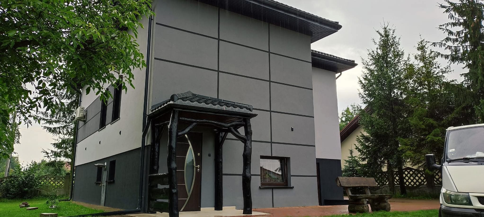 Dwukondygnacyjny dom z szarą elewacją, podzieloną czarnymi liniami, ozdobiony drewnianym zadaszeniem nad wejściem, otoczony zielenią.