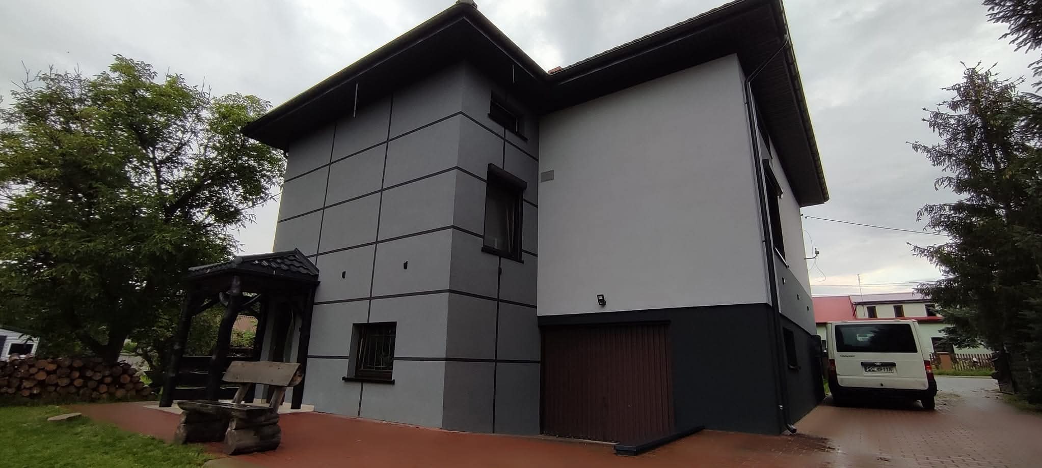 Dwukondygnacyjny dom z szarą i białą elewacją, ciemnymi akcentami i brązową bramą garażową. Widoczny fragment ogrodu i zaparkowany biały samochód.