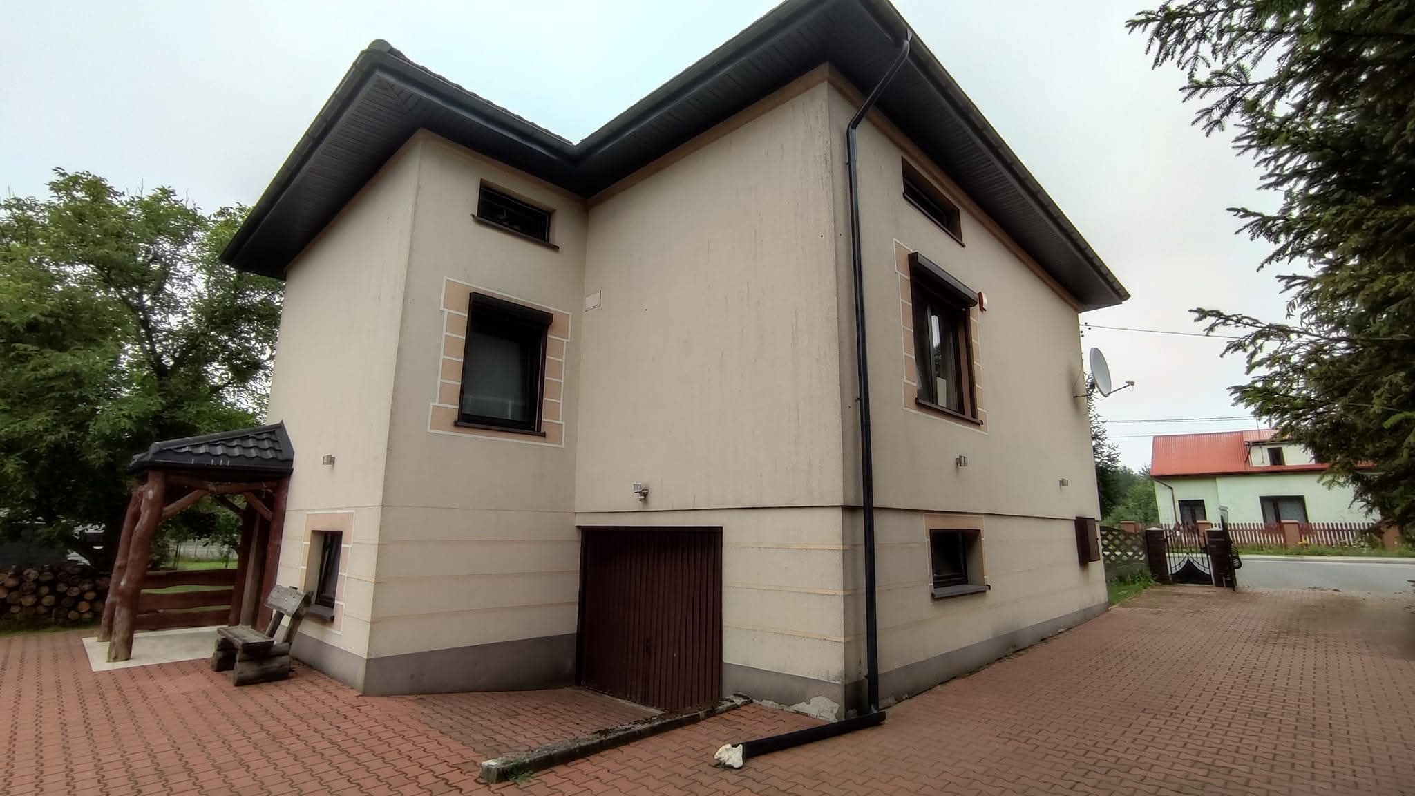Dwupiętrowy dom z beżową elewacją, czarnymi oknami i ciemnobrązową bramą garażową. Widoczne elementy dekoracyjne wokół okien i drewniana altana z boku budynku.