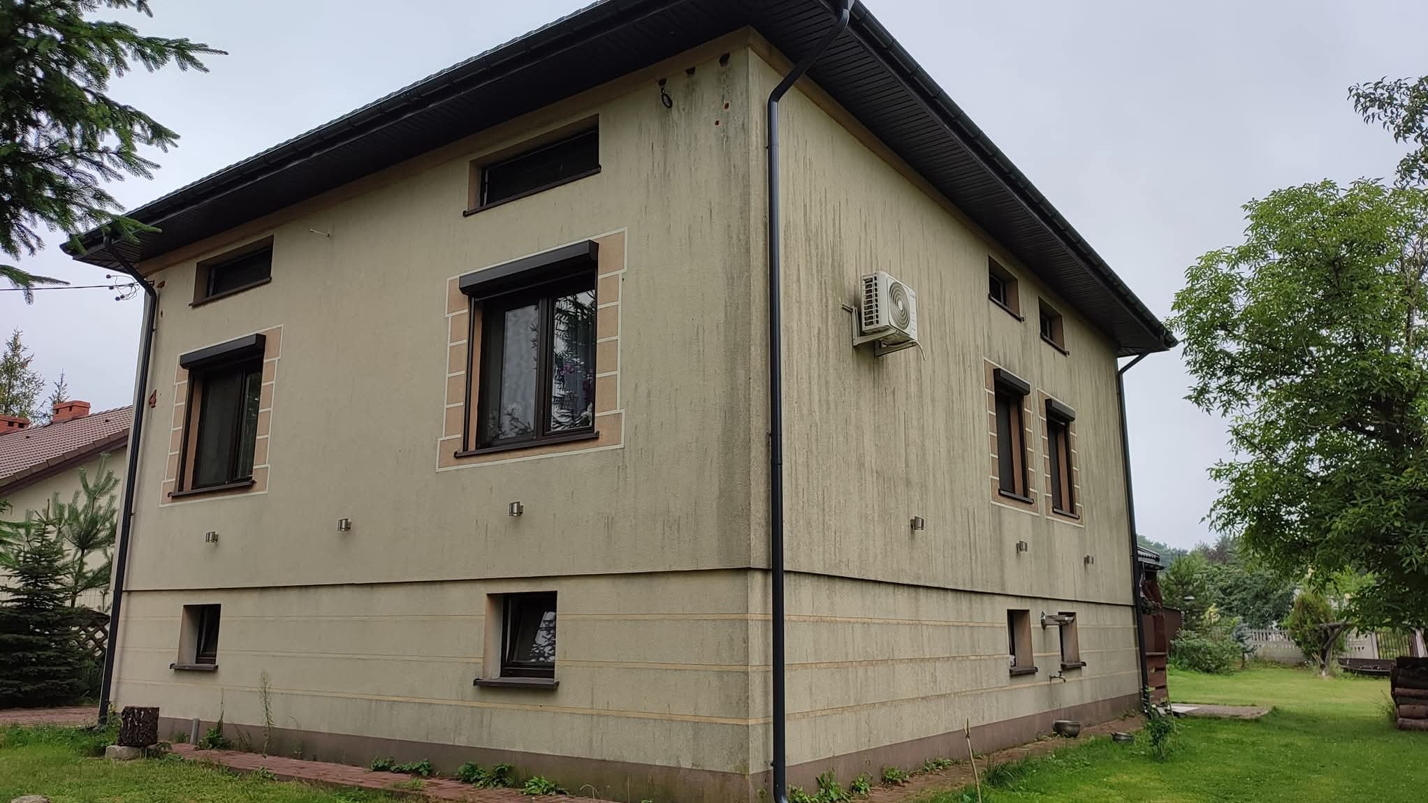 Dwukondygnacyjny dom z beżową elewacją, brązowymi ramami okiennymi i roletami. Widoczny klimatyzator na ścianie i fragment ogrodu. Ujęcie z perspektywy narożnika budynku.