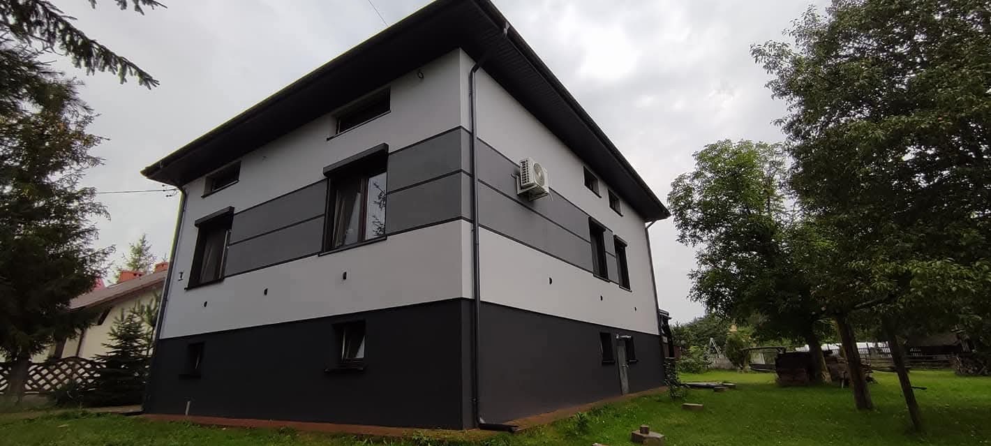 Dwukondygnacyjny dom z szarą elewacją w pasy i czarnym cokołem, klimatyzator na ścianie, widok z zewnątrz, zieleń w tle, pochmurne niebo.