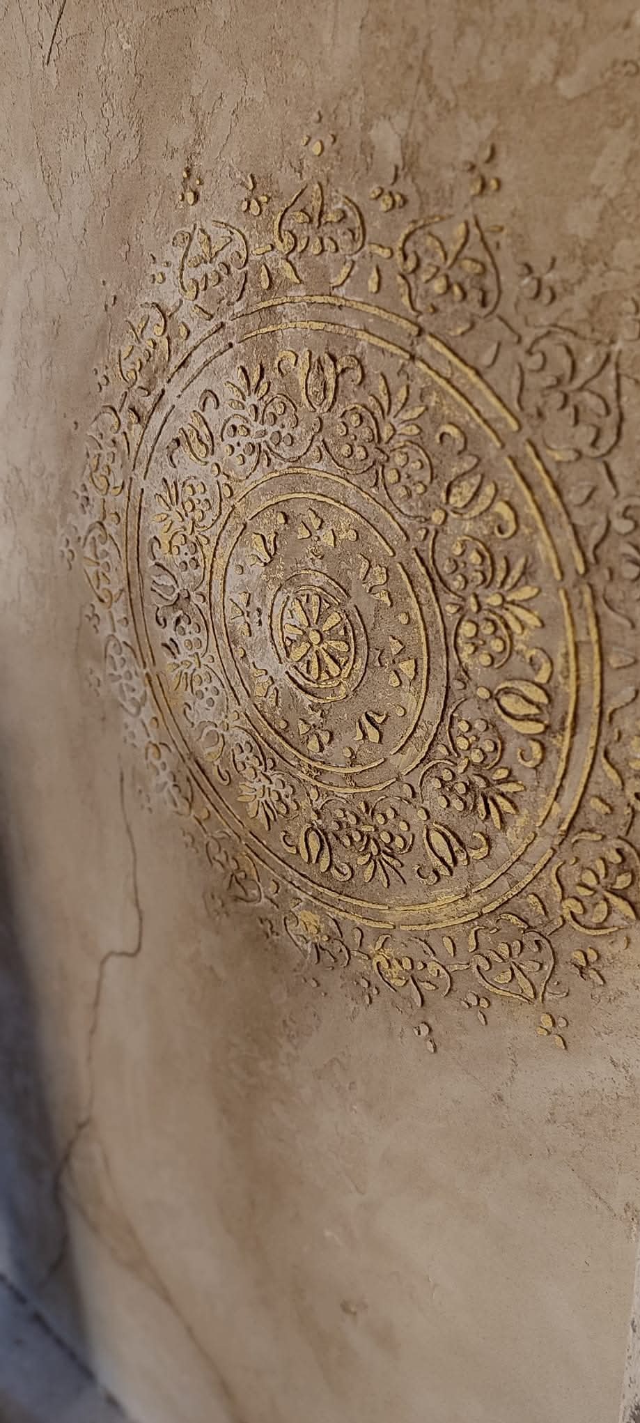 Dekoracyjna, wypukła rozeta z motywami roślinnymi, pomalowana złotą farbą, na beżowej ścianie z widoczną fakturą. Ujęcie z boku, podkreślające trójwymiarowość.