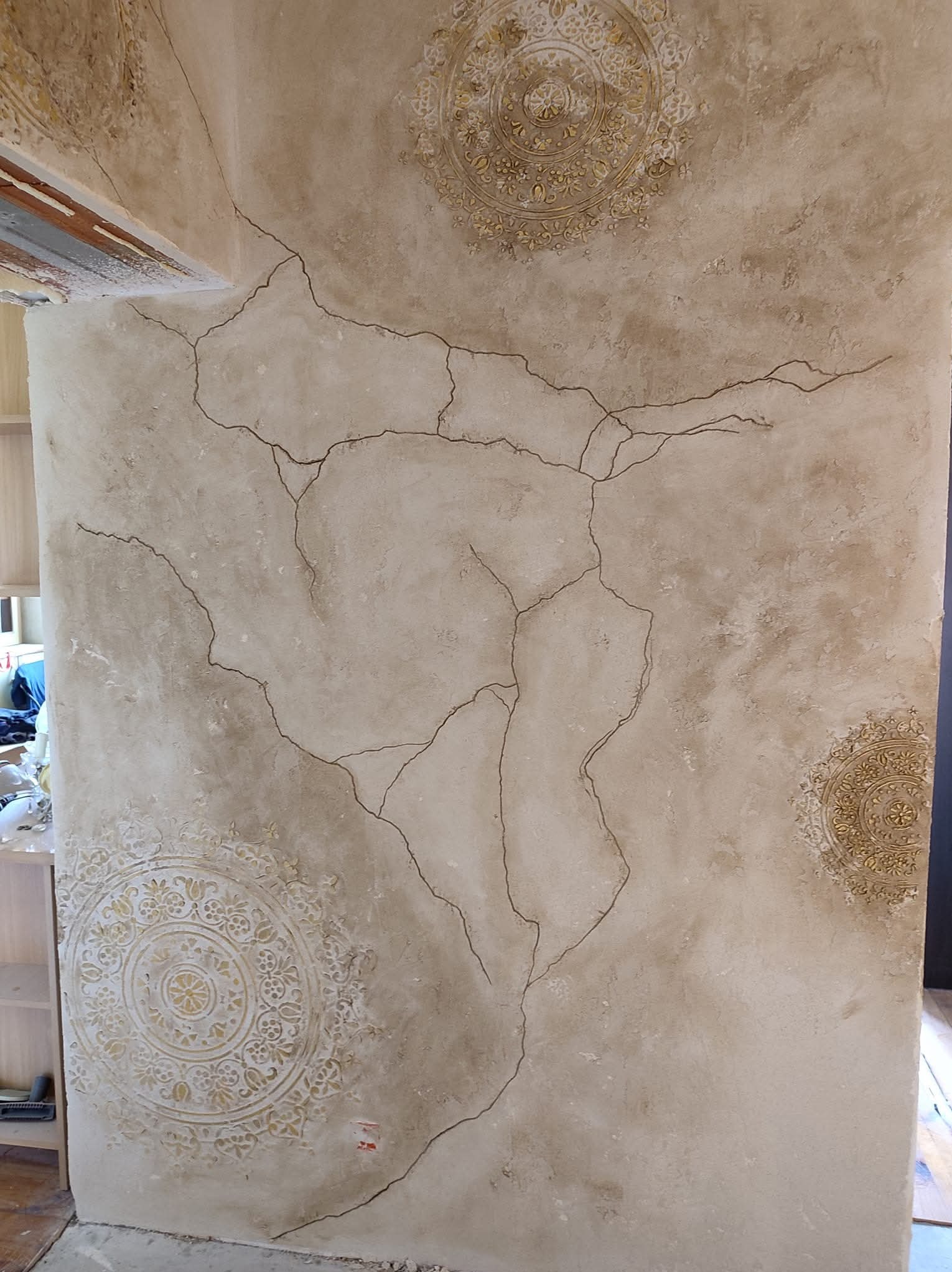 Dekoracyjna ściana z efektem spękań i ornamentami w odcieniach beżu i złota, widoczna część regału i fragment drewnianej belki stropowej.