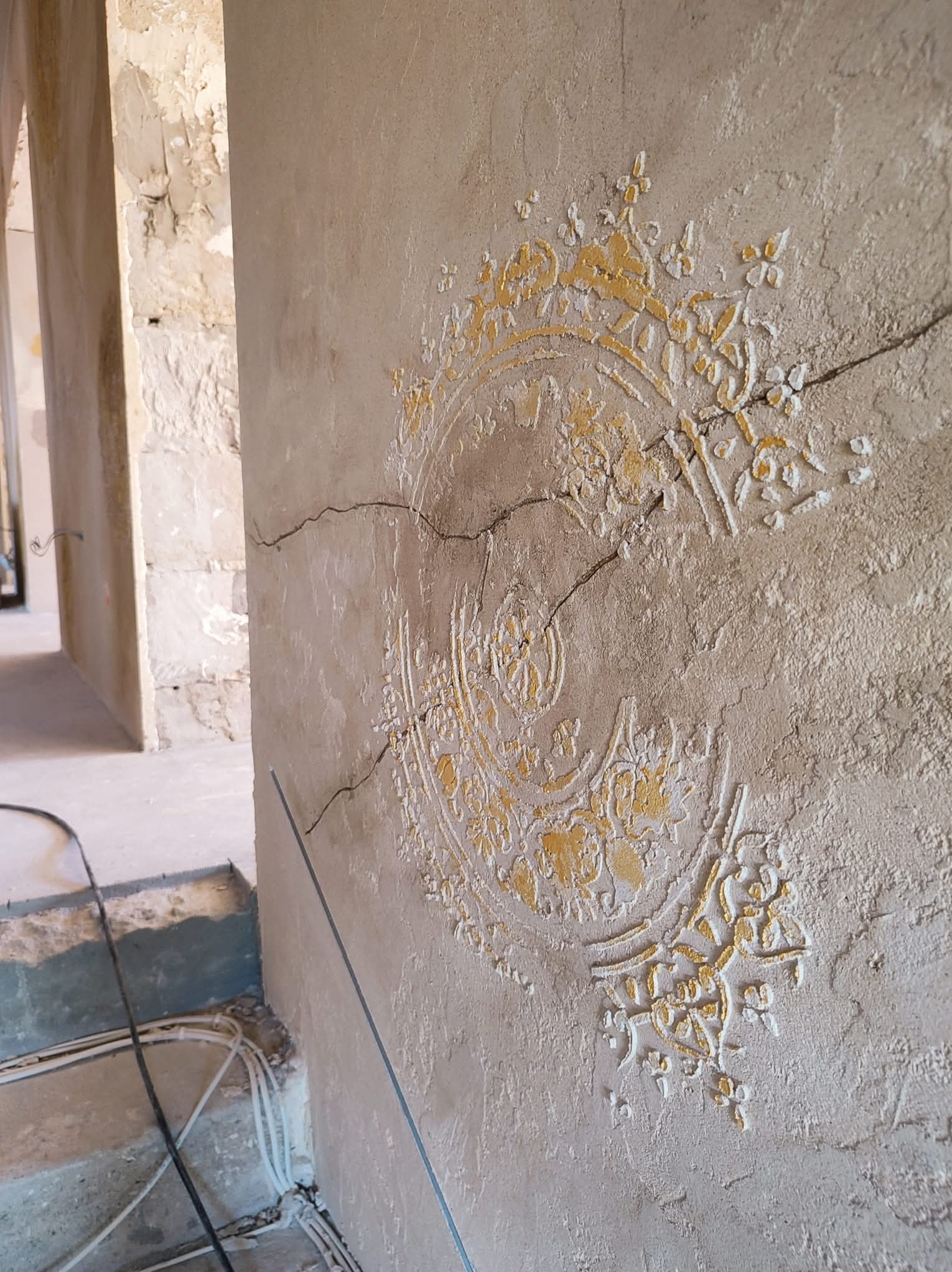 Ściana w trakcie szpachlowania z dekoracyjnym, częściowo pomalowanym wzorem. Widoczne pęknięcia i niedoskonałości, w tle surowy stan wnętrza.