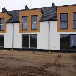 Nowoczesne segmenty mieszkalne w trakcie budowy, z białą i brązową elewacją, czarną dachówką i ramami okiennymi, widoczne żółte rury na tle błotnistego terenu.