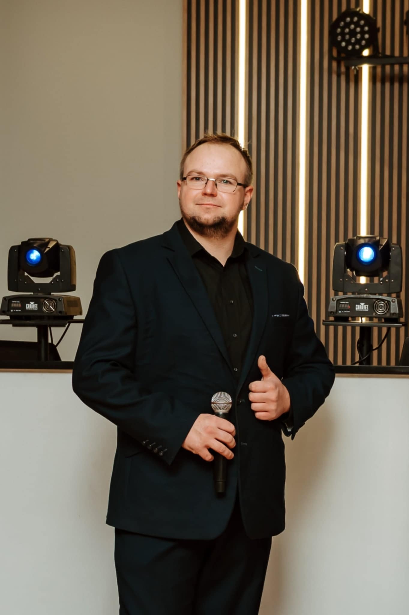Uśmiechnięty mężczyzna w garniturze trzyma mikrofon, w tle dwa reflektory sceniczne Chauvet i dekoracyjna ściana z pionowych paneli. Prezentacja sprzętu nagłośnieniowego.