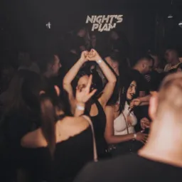 Rozbawiony tłum w klubie nocnym z neonowym napisem 'NIGHT'S PLAN' w tle, kobiety tańczą z podniesionymi rękami, atmosfera zabawy.