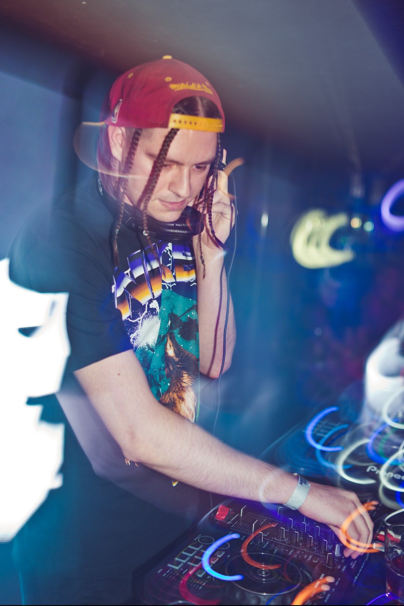 DJ z warkoczykami w czerwonej czapce, miksujący muzykę na konsolecie Pioneer DJ w klubie z rozmytymi neonami w tle.