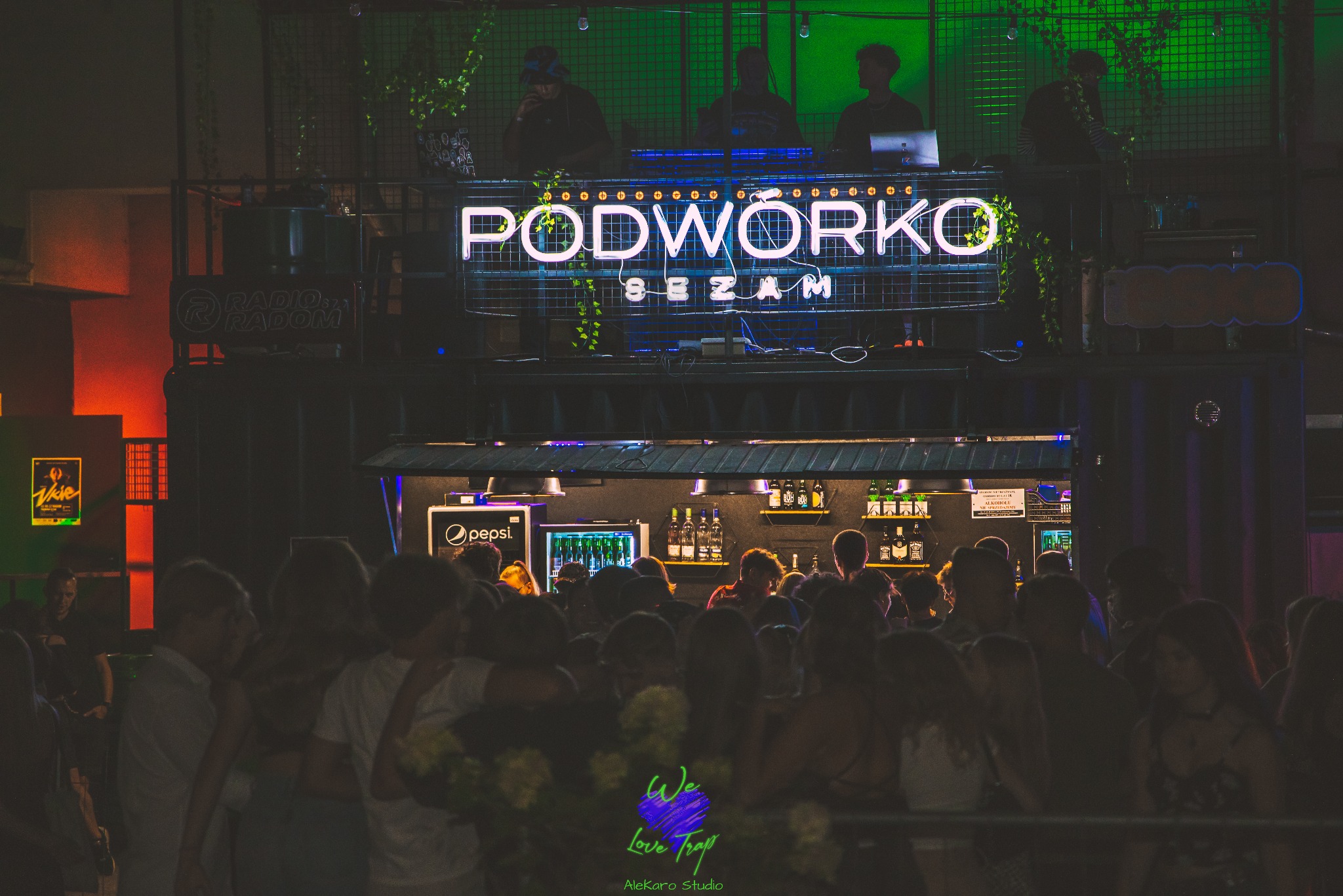 Tłum ludzi przed barem 'Podwórko Sezam' podczas imprezy z DJ-ami grającymi muzykę na górze, oświetlenie neonowe i dekoracje roślinne.