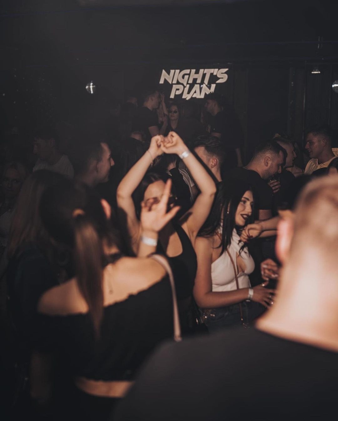 Rozbawiony tłum w klubie nocnym z neonowym napisem 'NIGHT'S PLAN' w tle, kobiety tańczą z podniesionymi rękami, atmosfera zabawy.