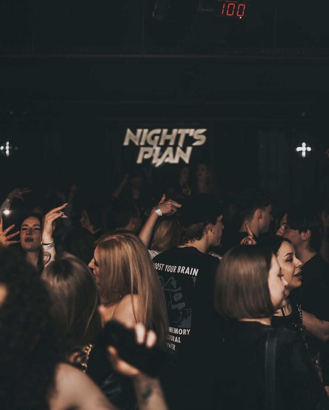 Tłum ludzi w ciemnym klubie nocnym, z podświetlonym napisem 'NIGHT'S PLAN' w tle i czerwonym wyświetlaczem '100' nad nim.