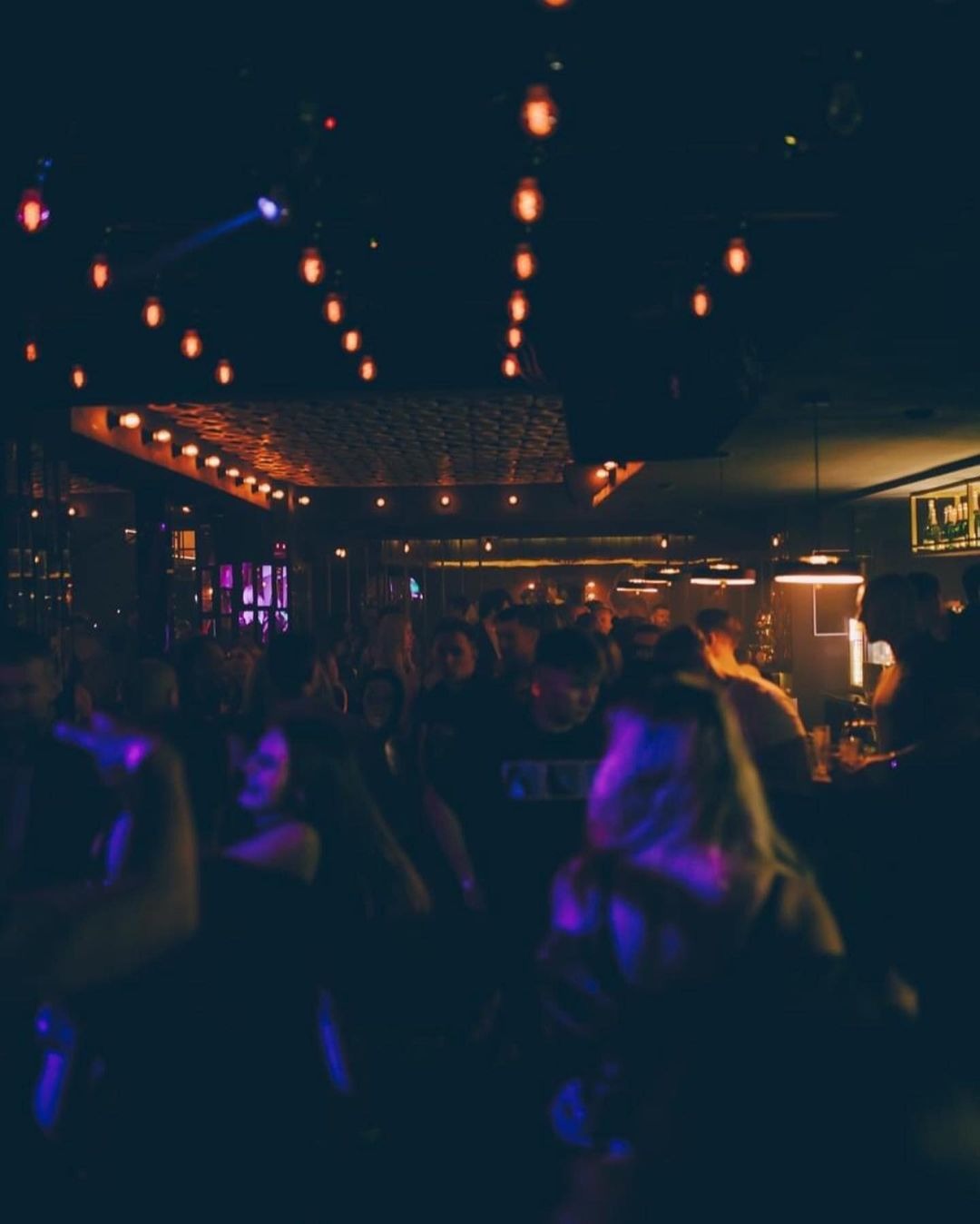 Zabawa w klubie nocnym z przyciemnionym oświetleniem, widoczne tańczące osoby i dekoracyjne żarówki na suficie.