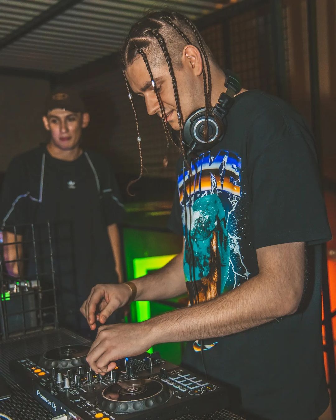 DJ z warkoczykami i słuchawkami na szyi miksuje muzykę na profesjonalnym sprzęcie Pioneer DJ, w tle postać w czarnej bluzie z czapką.