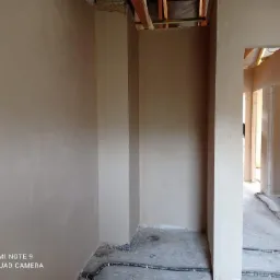 Wnętrze w trakcie prac wykończeniowych: świeżo nałożone, gładkie tynki w odcieniach beżu na ścianach i filarach, widoczne instalacje elektryczne na surowej podłodze, sufit w stanie deweloperskim...