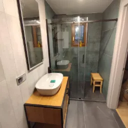 Nowoczesna łazienka z umywalką nablatową na drewnianej szafce z czarnymi elementami, kabiną prysznicową typu walk-in z przesuwnymi drzwiami i drewnianym stołkiem wewnątrz, oraz dużym lustrem...