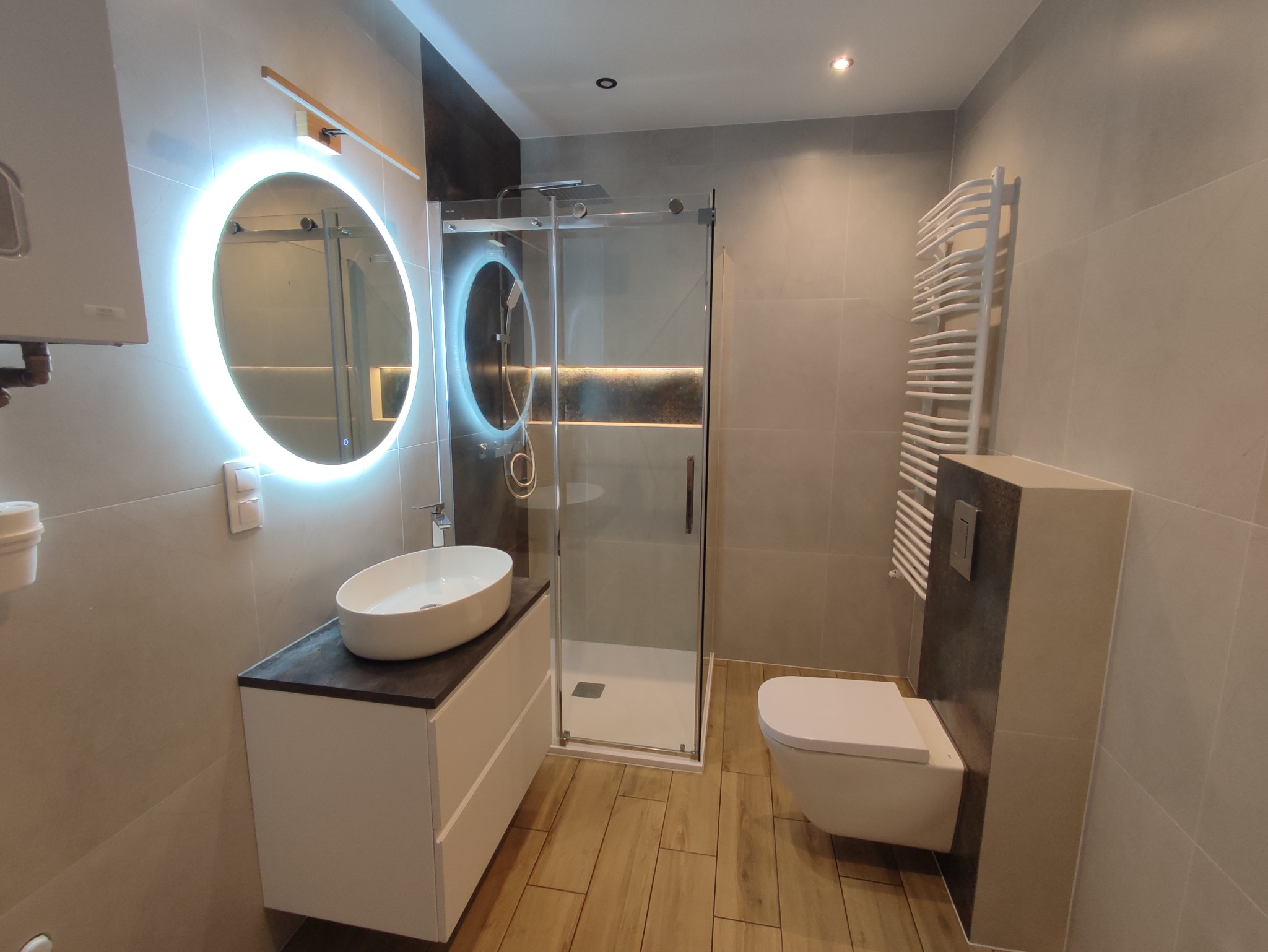 Nowoczesna łazienka z drewnopodobną podłogą, umywalką nablatową na białej szafce z ciemnym blatem, okrągłym lustrem z podświetleniem LED, kabiną prysznicową ze szklanymi drzwiami i wiszącą toaletą.