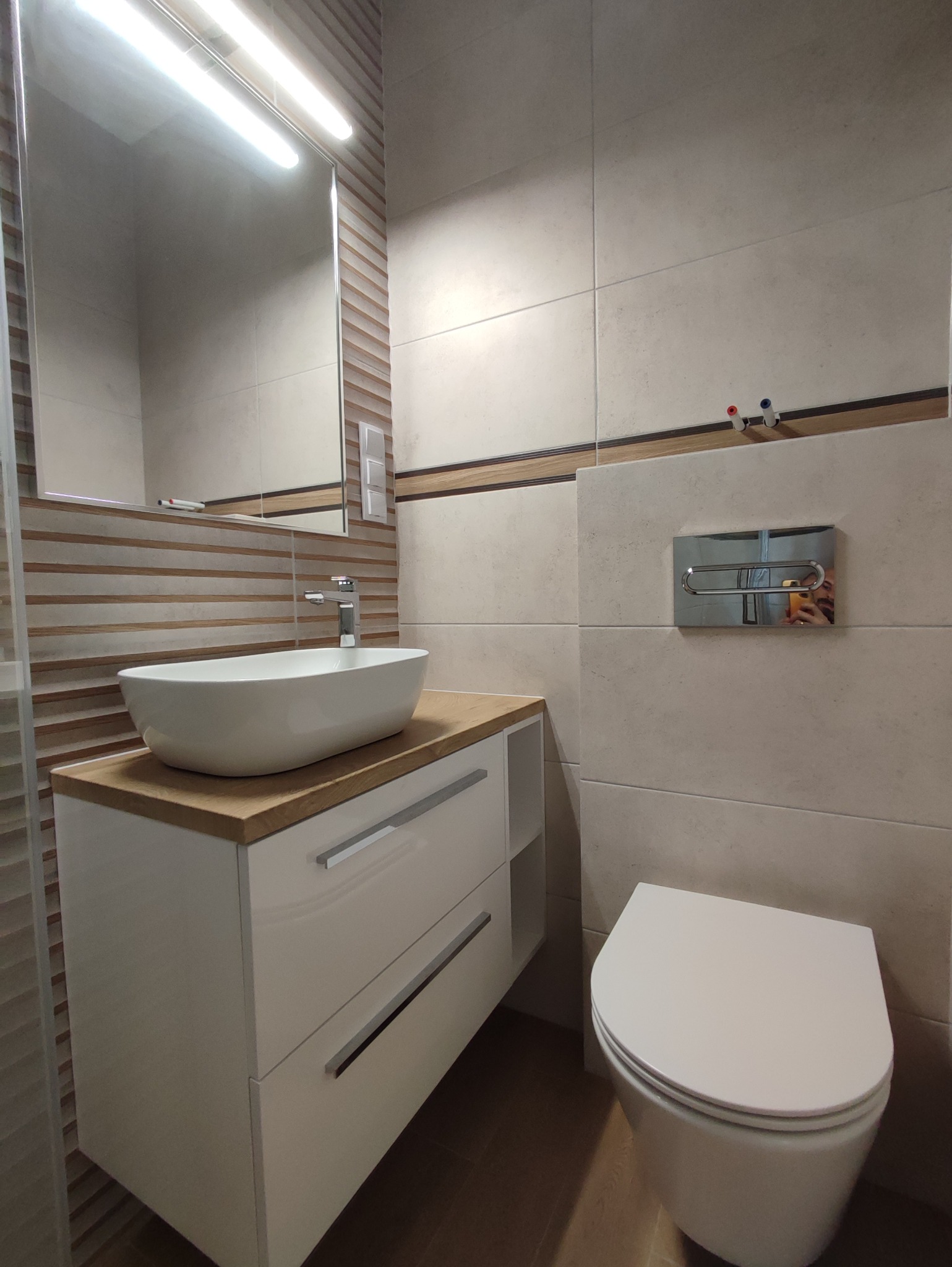 Nowoczesna łazienka z umywalką nablatową na szafce z szufladami, podświetlanym lustrem i wiszącą toaletą, wykończona płytkami w odcieniach beżu i brązu z dekoracyjnym pasem.