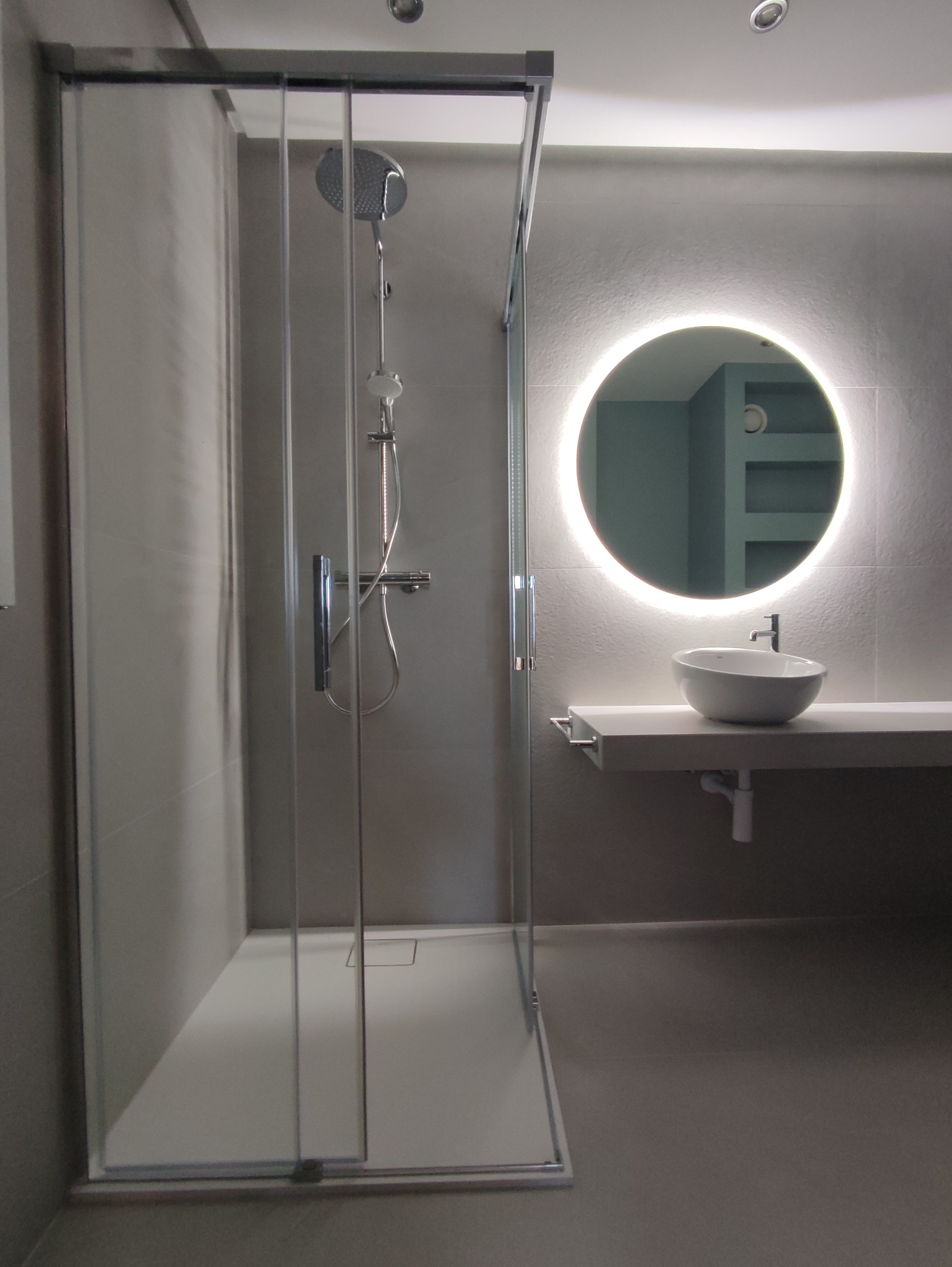 Nowoczesna łazienka w odcieniach szarości z kabiną prysznicową typu walk-in z przezroczystego szkła i okrągłym lustrem z podświetleniem LED nad umywalką nablatową.
