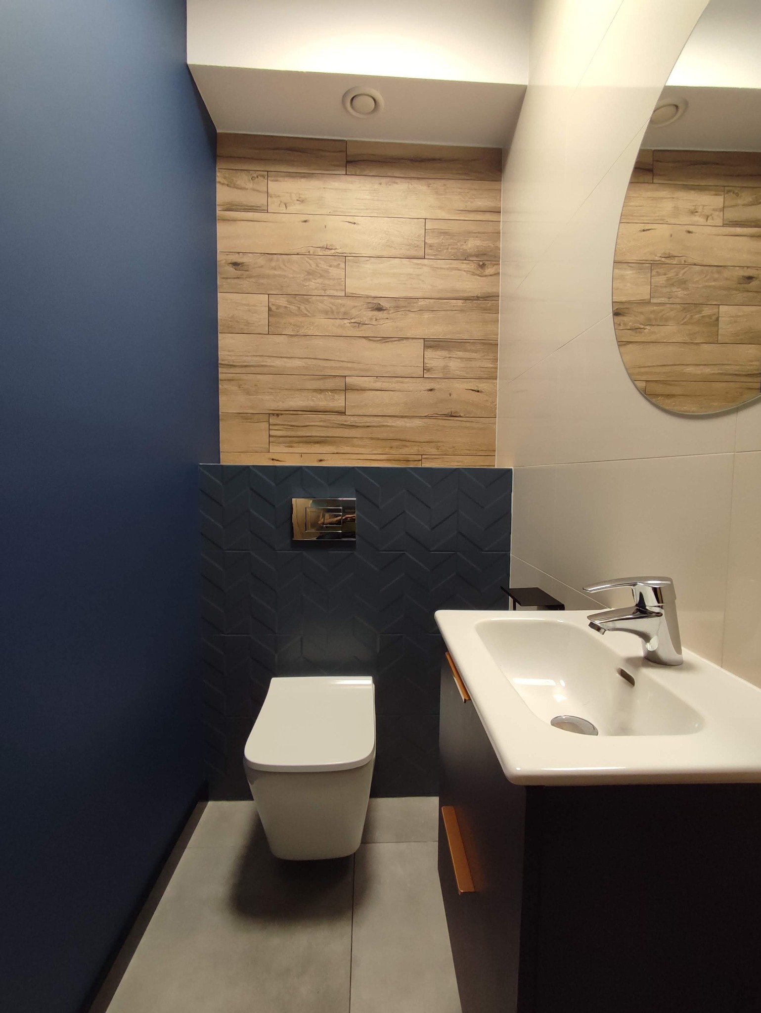 Nowoczesna łazienka z toaletą podwieszaną, umywalką nablatową na szafce w kolorze grafitowym, ściana pomalowana na granatowo, oraz płytki imitujące drewno na górnej części ściany i ciemne płytki...
