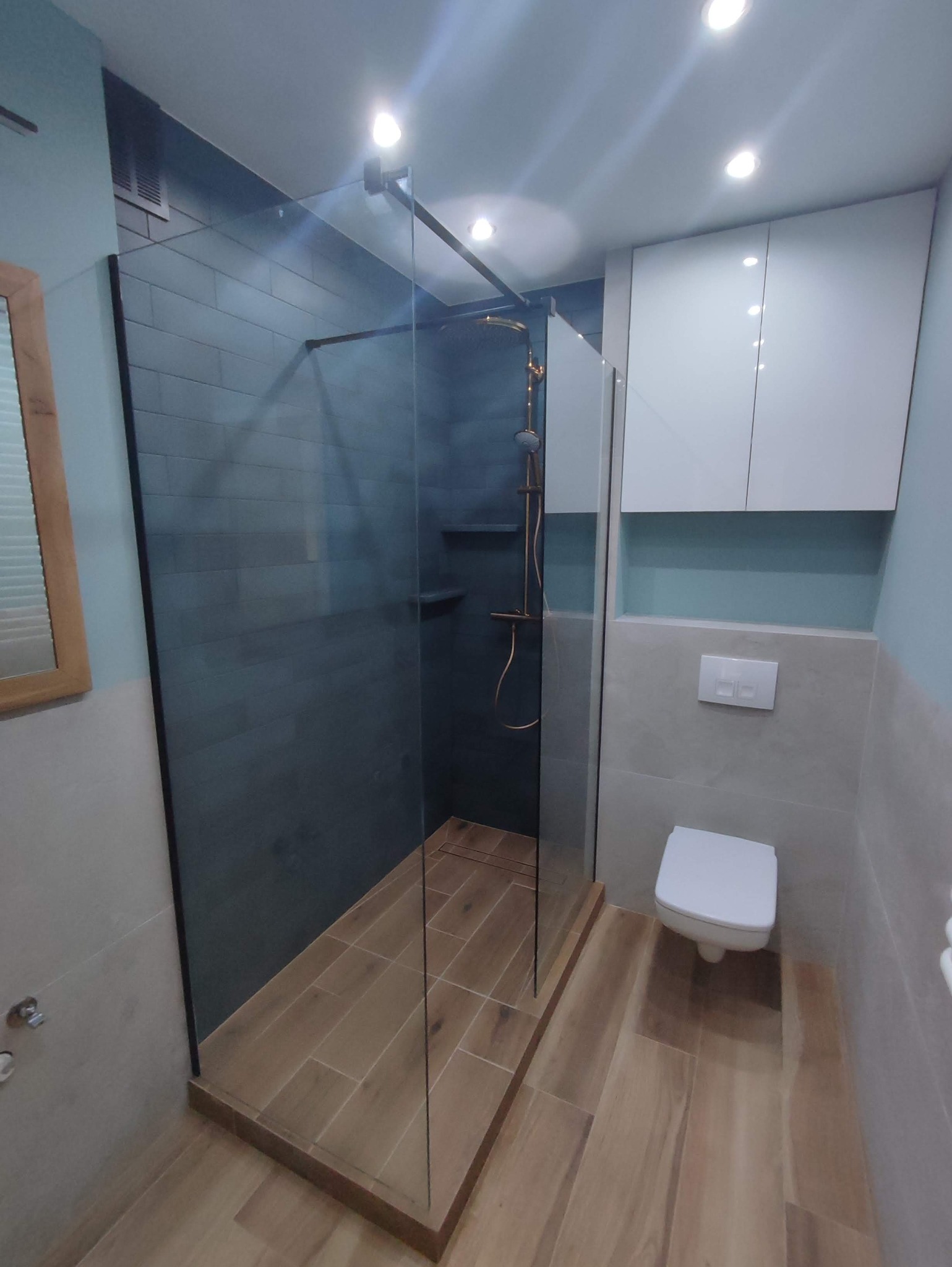 Nowoczesna łazienka z kabiną prysznicową typu walk-in z grafitowymi płytkami, złotą armaturą i drewnopodobną podłogą, oświetlona punktowymi lampami sufitowymi.