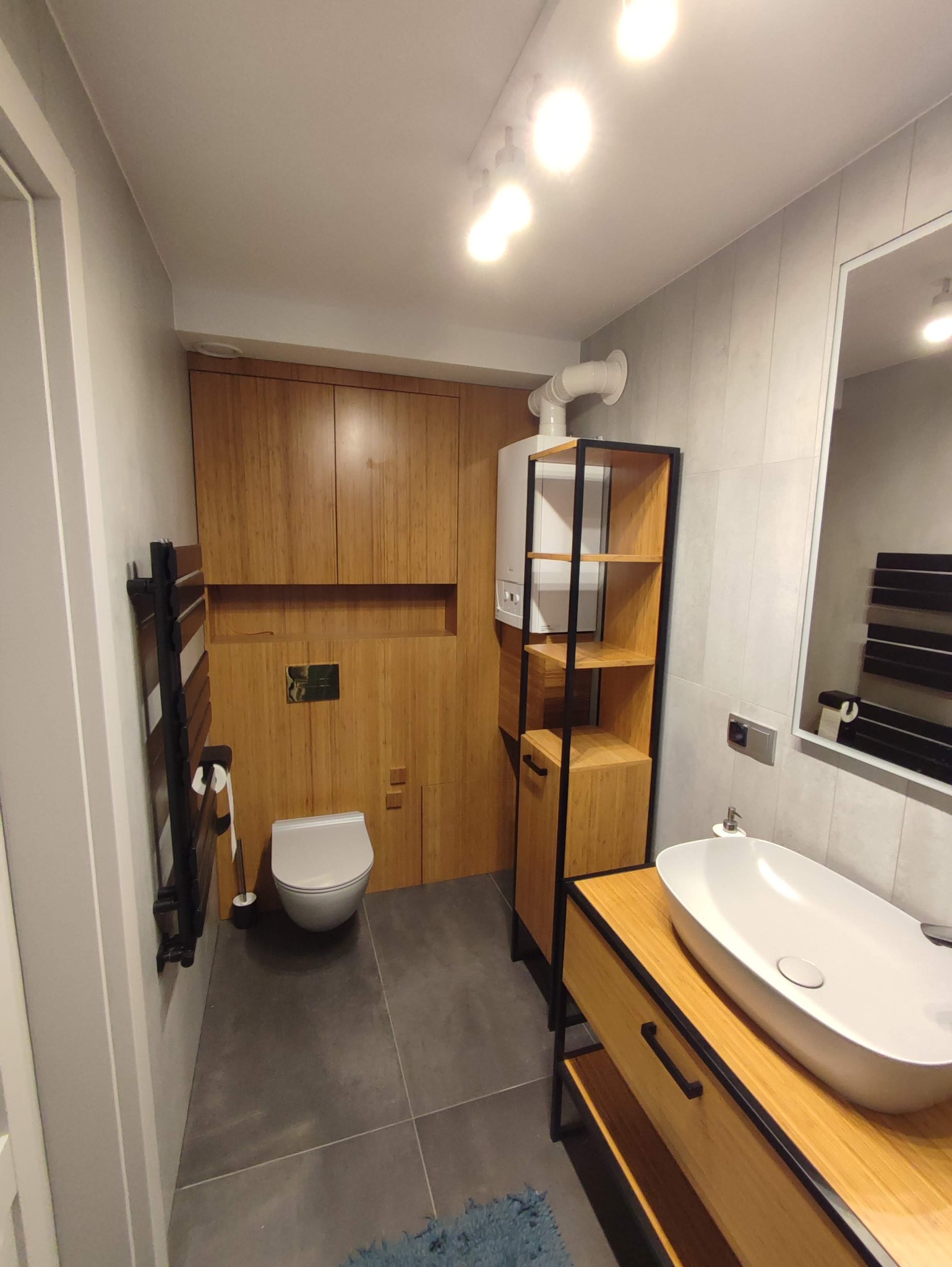 Nowoczesna łazienka z drewnianymi akcentami: zabudowa z bambusowych paneli, podwieszana toaleta, umywalka nablatowa na szafce z otwartymi półkami, grzejnik drabinkowy i piec gazowy w zabudowie.