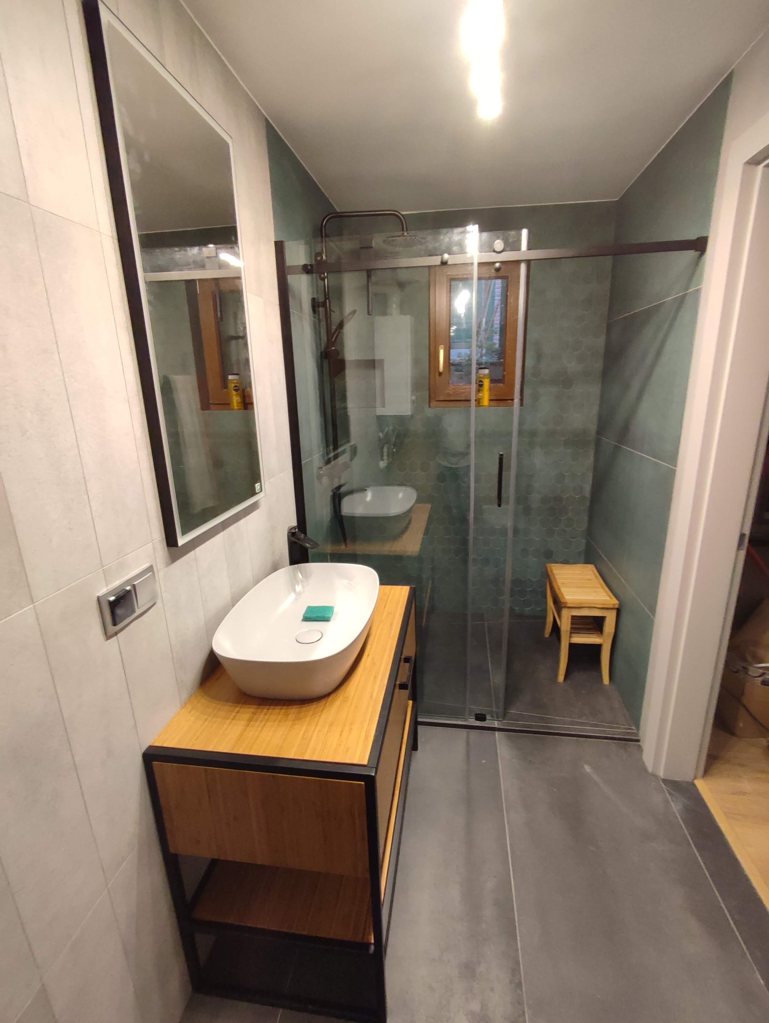 Nowoczesna łazienka z umywalką nablatową na drewnianej szafce z czarnymi elementami, kabiną prysznicową typu walk-in z przesuwnymi drzwiami i drewnianym stołkiem wewnątrz, oraz dużym lustrem...