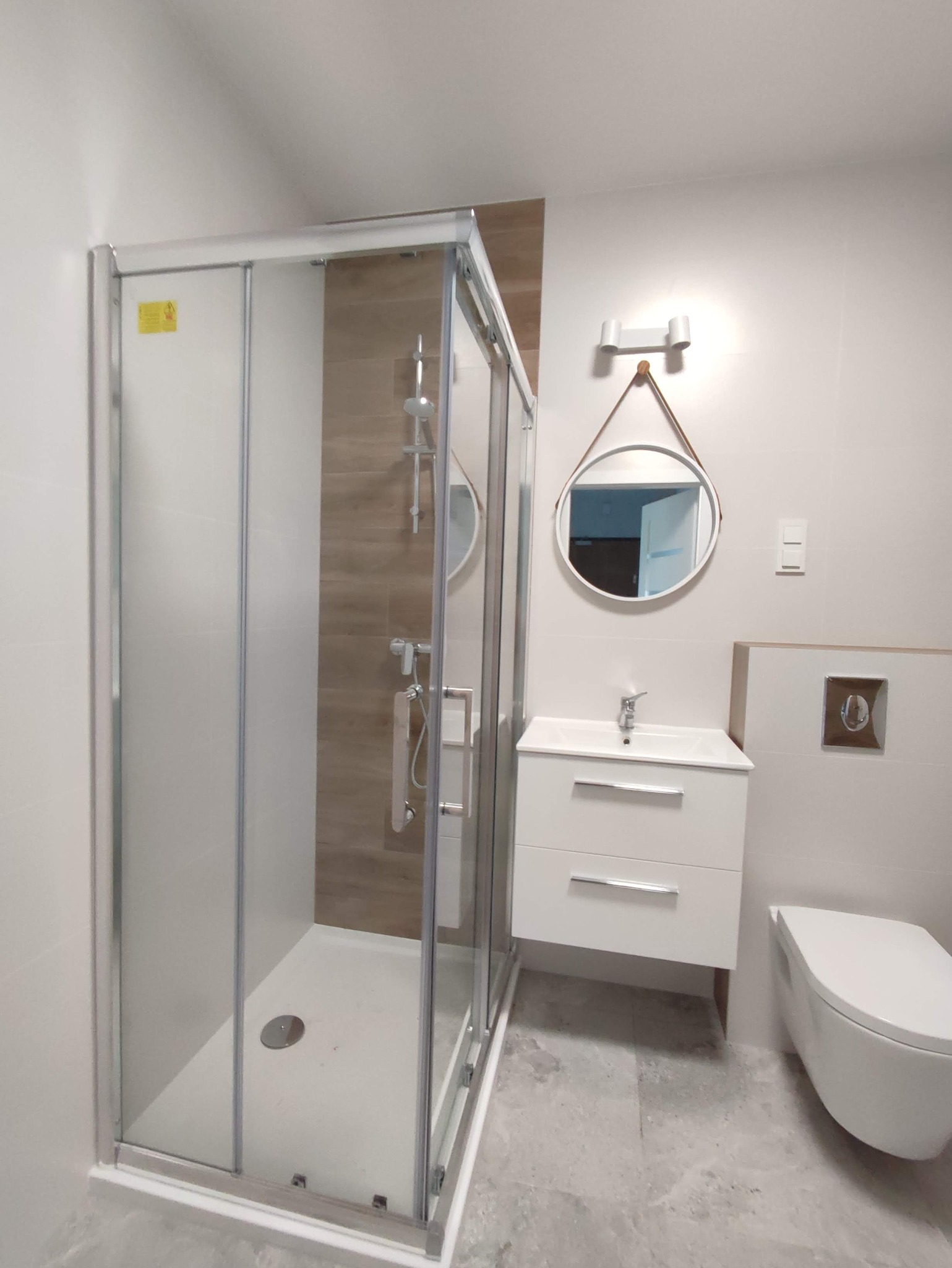 Nowoczesna łazienka po remoncie: kabina prysznicowa z przesuwnymi drzwiami, biała szafka z umywalką, okrągłe lustro z brązowym paskiem, podwieszana toaleta i szare płytki podłogowe.