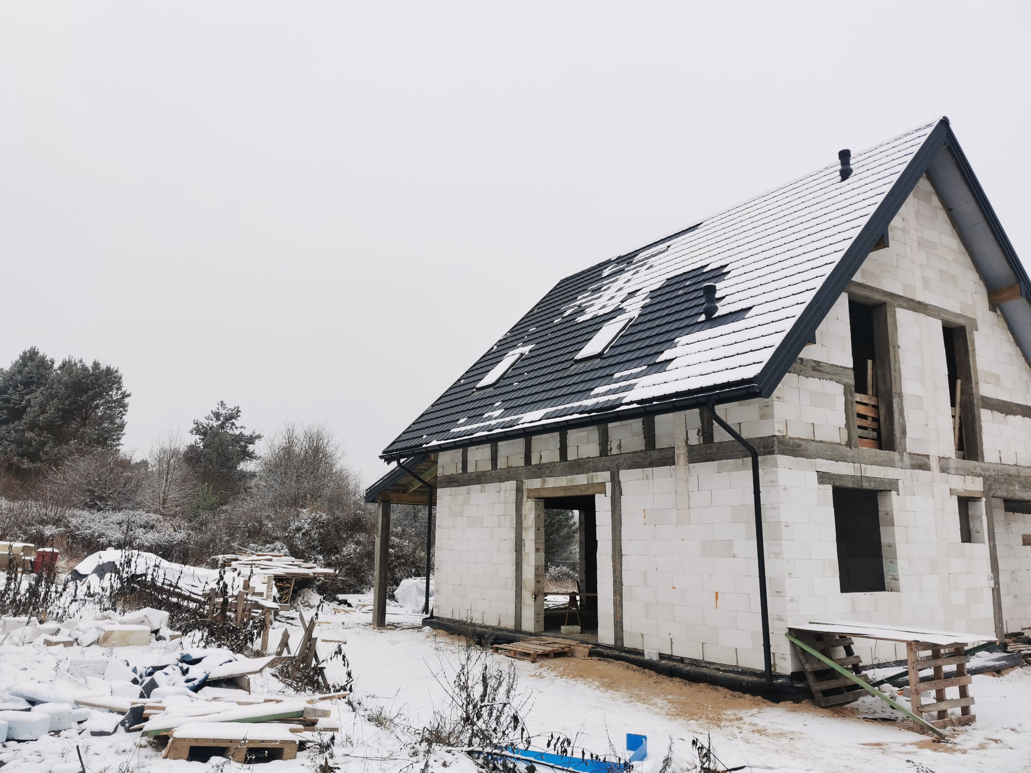 Dom w trakcie budowy, częściowo pokryty ciemną dachówką, zimowy krajobraz, widoczne pustaki i elementy konstrukcyjne.