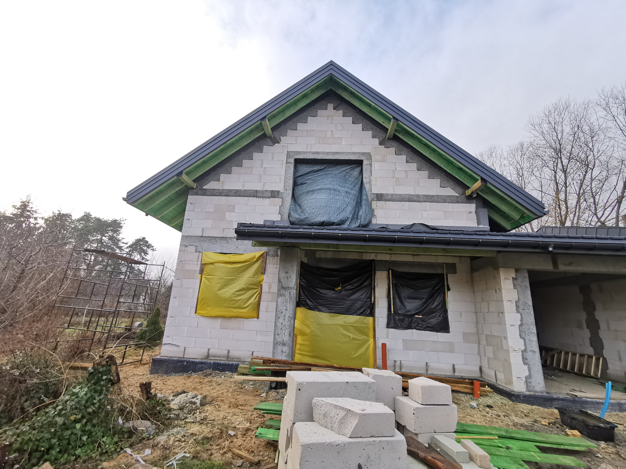 Dom w trakcie budowy z widocznym dachem z czarnej blachy trapezowej i elementami obróbki blacharskiej, okna zabezpieczone folią, a na pierwszym planie ułożone bloczki betonowe.