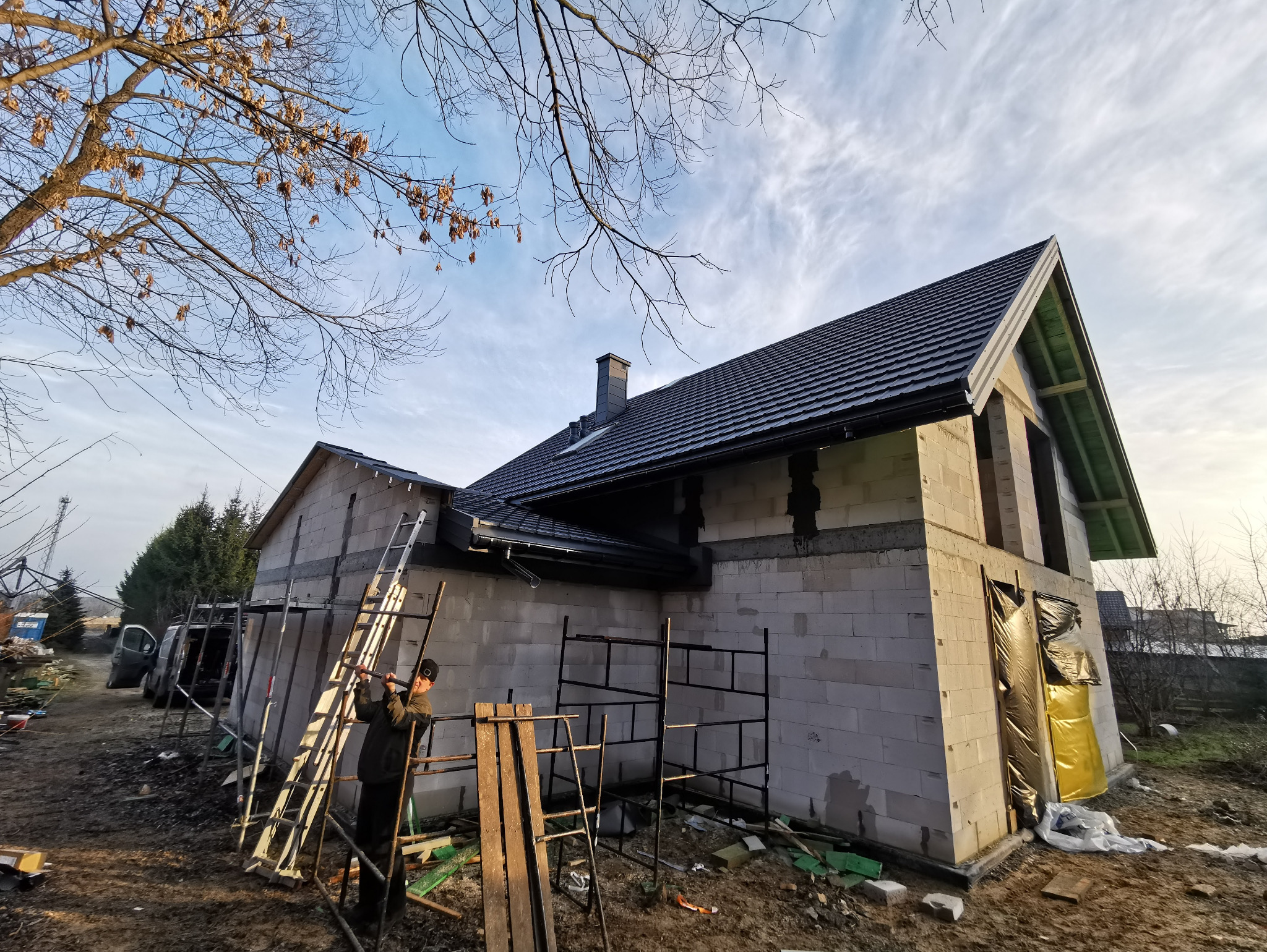 Budowa domu jednorodzinnego z ciemnym dachem, widoczne rusztowania i pracownik wspinający się po drabinie, w tle drzewa bez liści i pochmurne niebo.