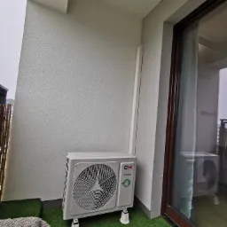 Zewnętrzna jednostka klimatyzacji CH Inverter zainstalowana na balkonie z sztuczną trawą, widoczna z bliska, z fragmentem szarego ręcznika w lewym dolnym rogu.