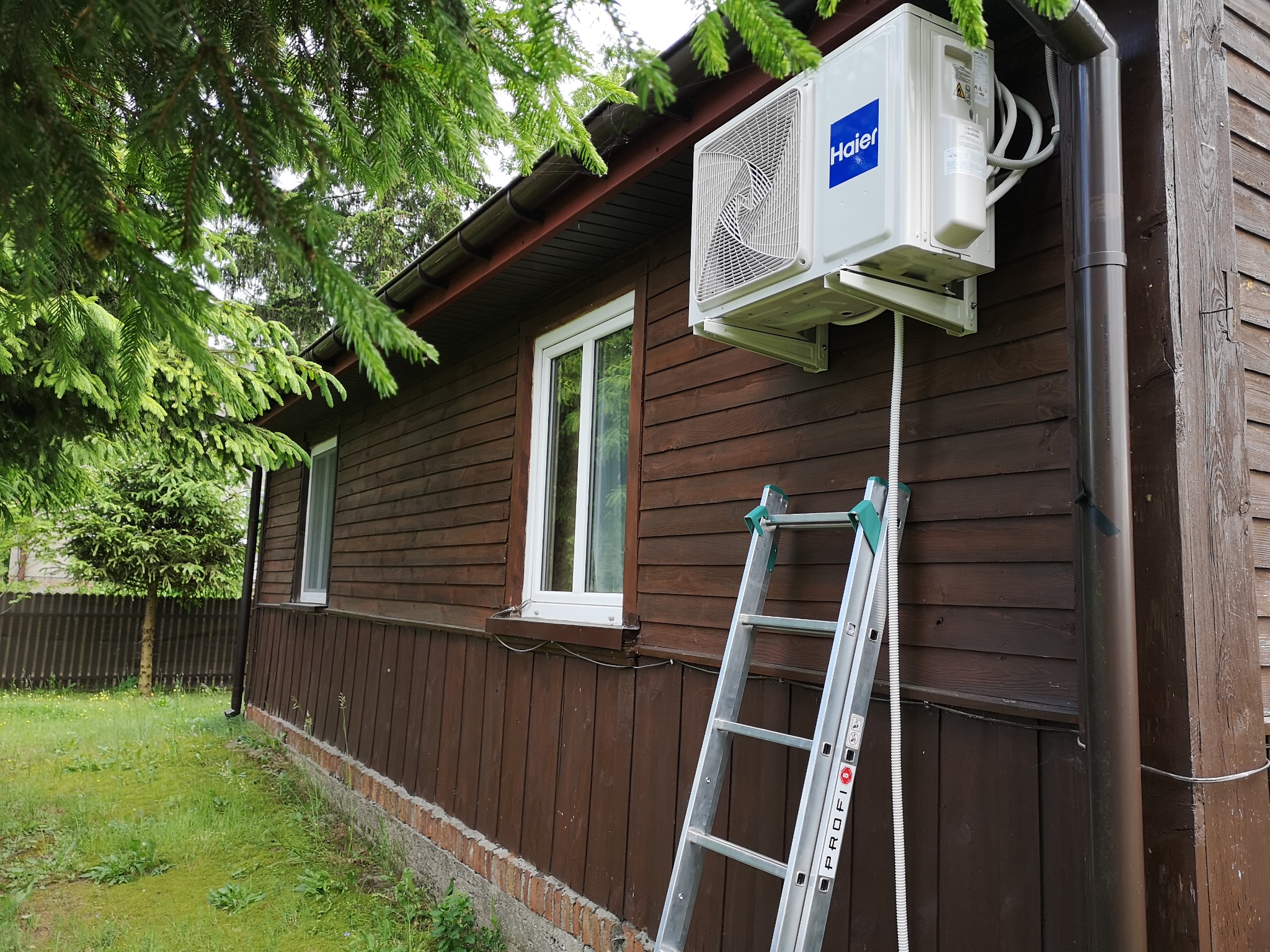 Zewnętrzna jednostka klimatyzatora marki Haier zamontowana na brązowej, drewnianej ścianie domu, widoczna drabina z napisem 'PROFI' i przewody prowadzące do urządzenia.