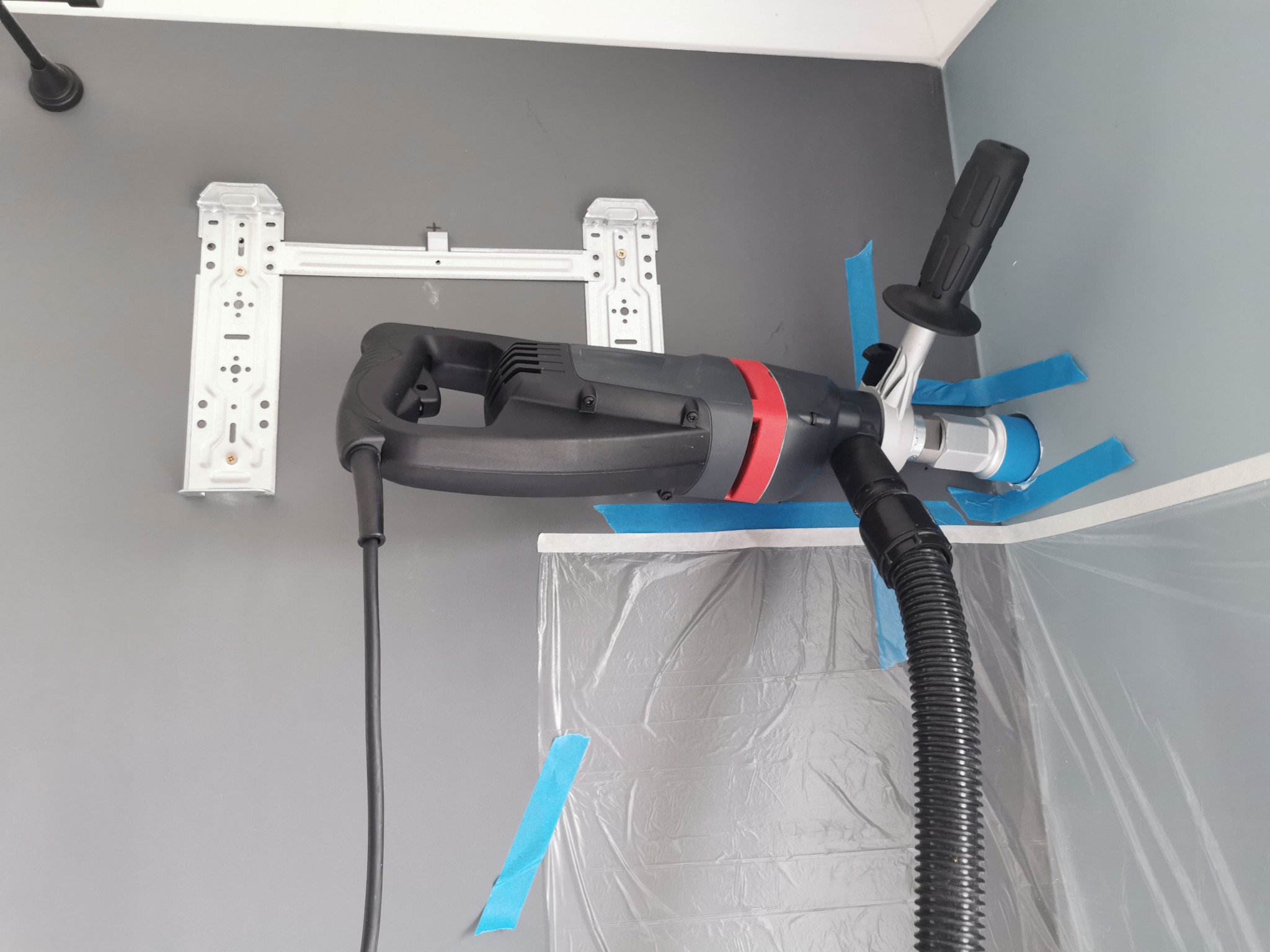 Wiertnica koronowa z odkurzaczem do wiercenia bezpyłowego, przymocowana taśmą malarską do ściany obok wspornika montażowego klimatyzatora, osłoniętej folią malarską