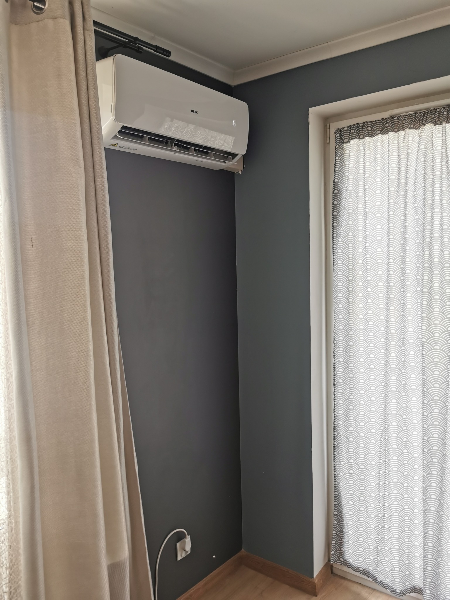 Biała jednostka wewnętrzna klimatyzatora marki AUX zamontowana na szarej ścianie w rogu pokoju, obok okna z zasłoną w japońskie wzory i beżową zasłoną.