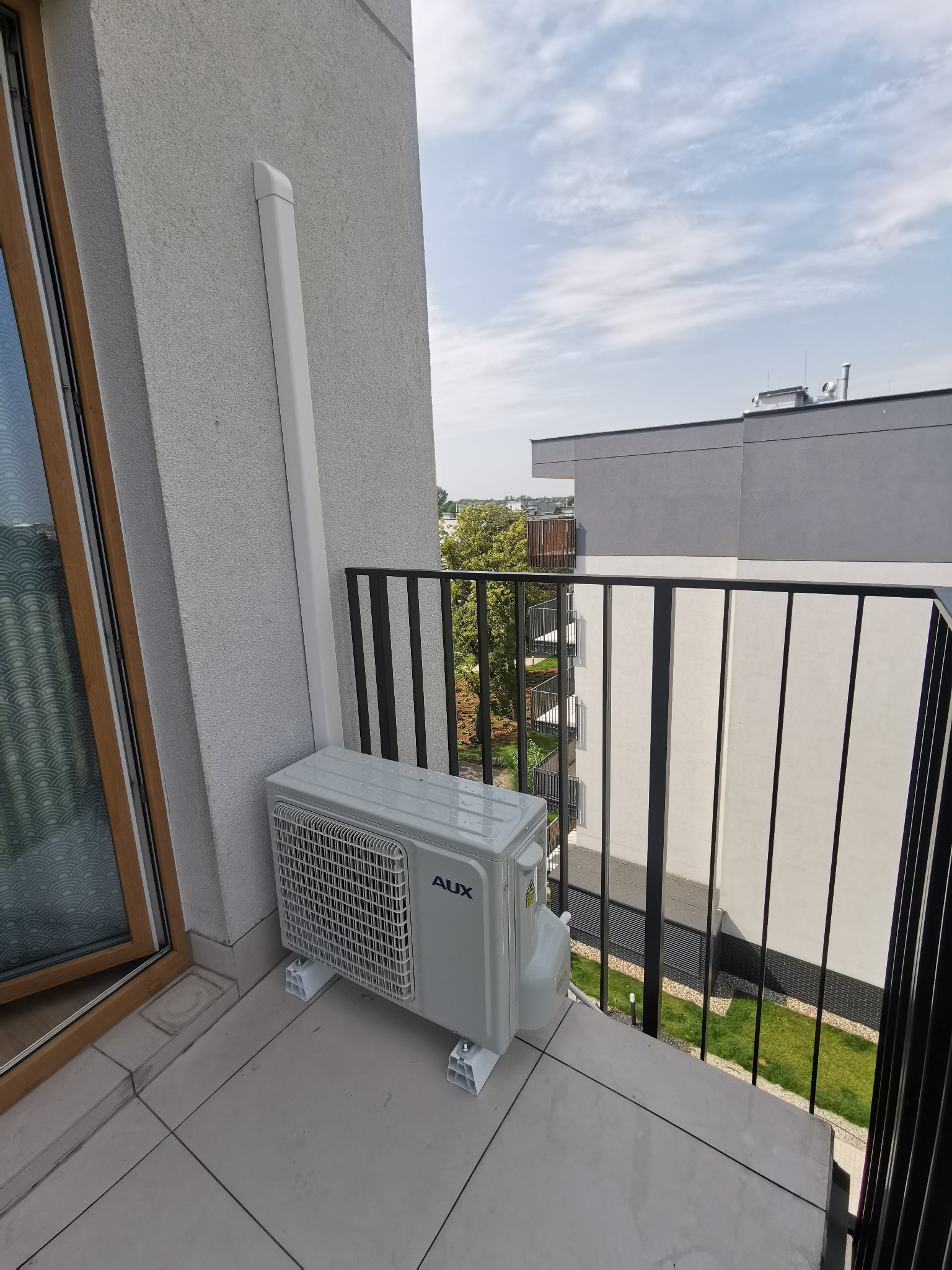 Zewnętrzna jednostka klimatyzacji AUX zamontowana na balkonie nowoczesnego apartamentu z widokiem na inne budynki i zieleń.