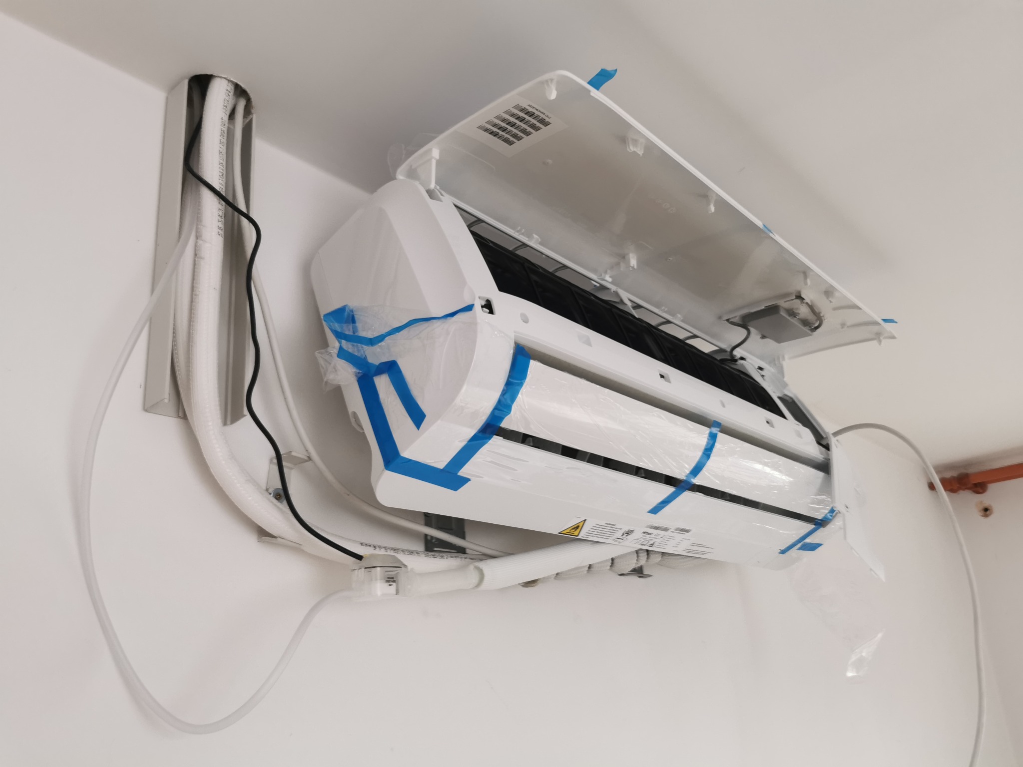 Świeżo zamontowany, biały klimatyzator typu split z otwartą obudową, zabezpieczony folią i niebieską taśmą, widoczne przewody w białej osłonie przymocowane do ściany.