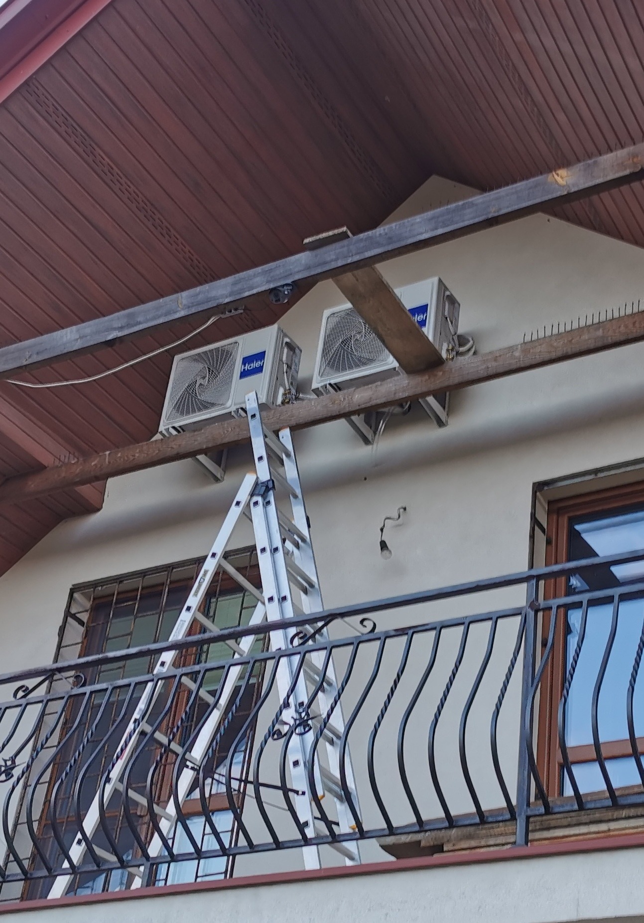 Dwa białe klimatyzatory marki Haier zamontowane na zewnątrz budynku, widoczne z balkonu, z rozstawioną aluminiową drabiną opartą o drewnianą belkę, zabezpieczoną kolcami przeciw ptakom.