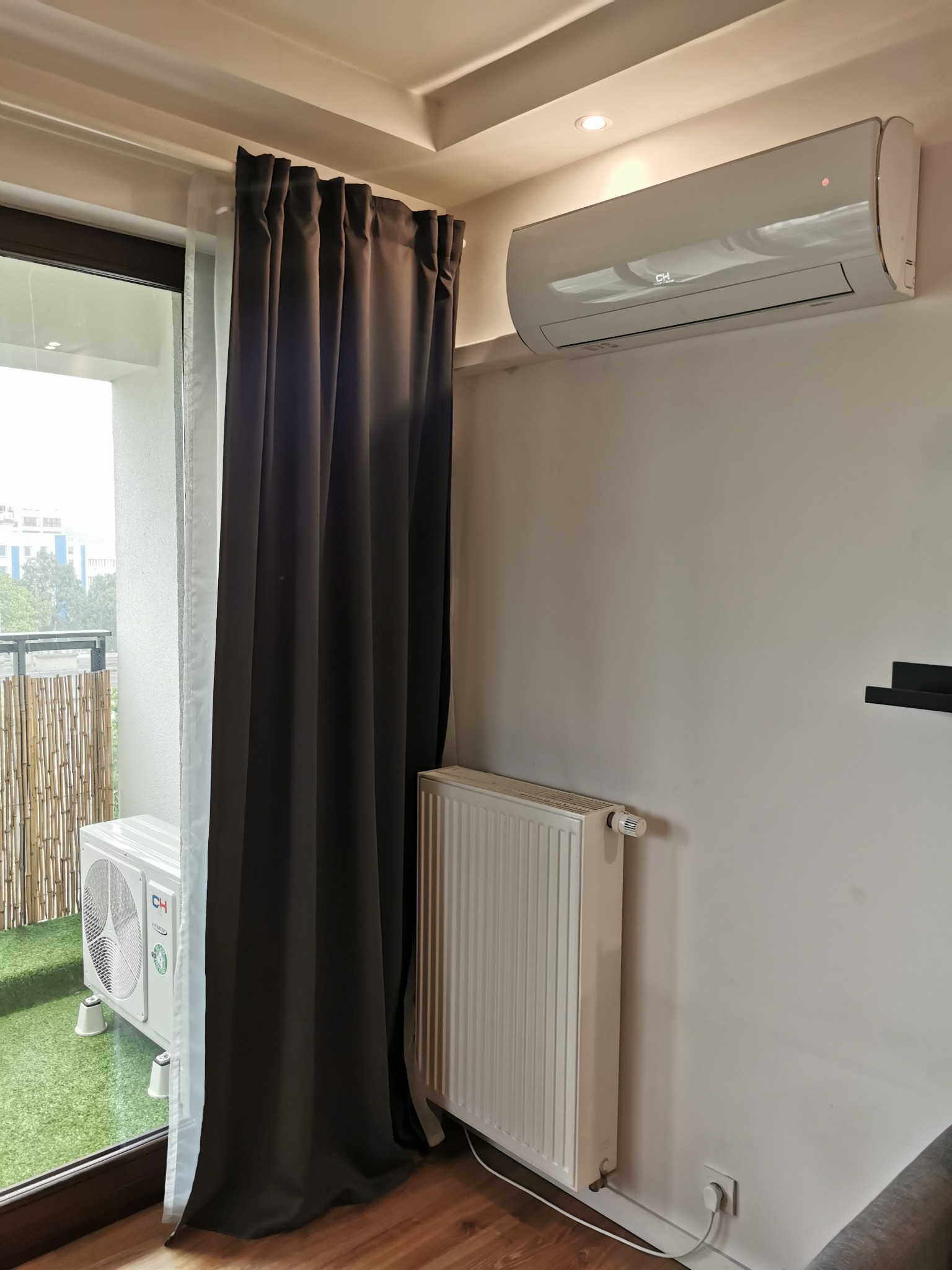 Biała jednostka wewnętrzna klimatyzatora zamontowana na ścianie w pokoju z grzejnikiem, widok na balkon z jednostką zewnętrzną klimatyzatora i sztuczną trawą, zasłonięty ciemnymi zasłonami.