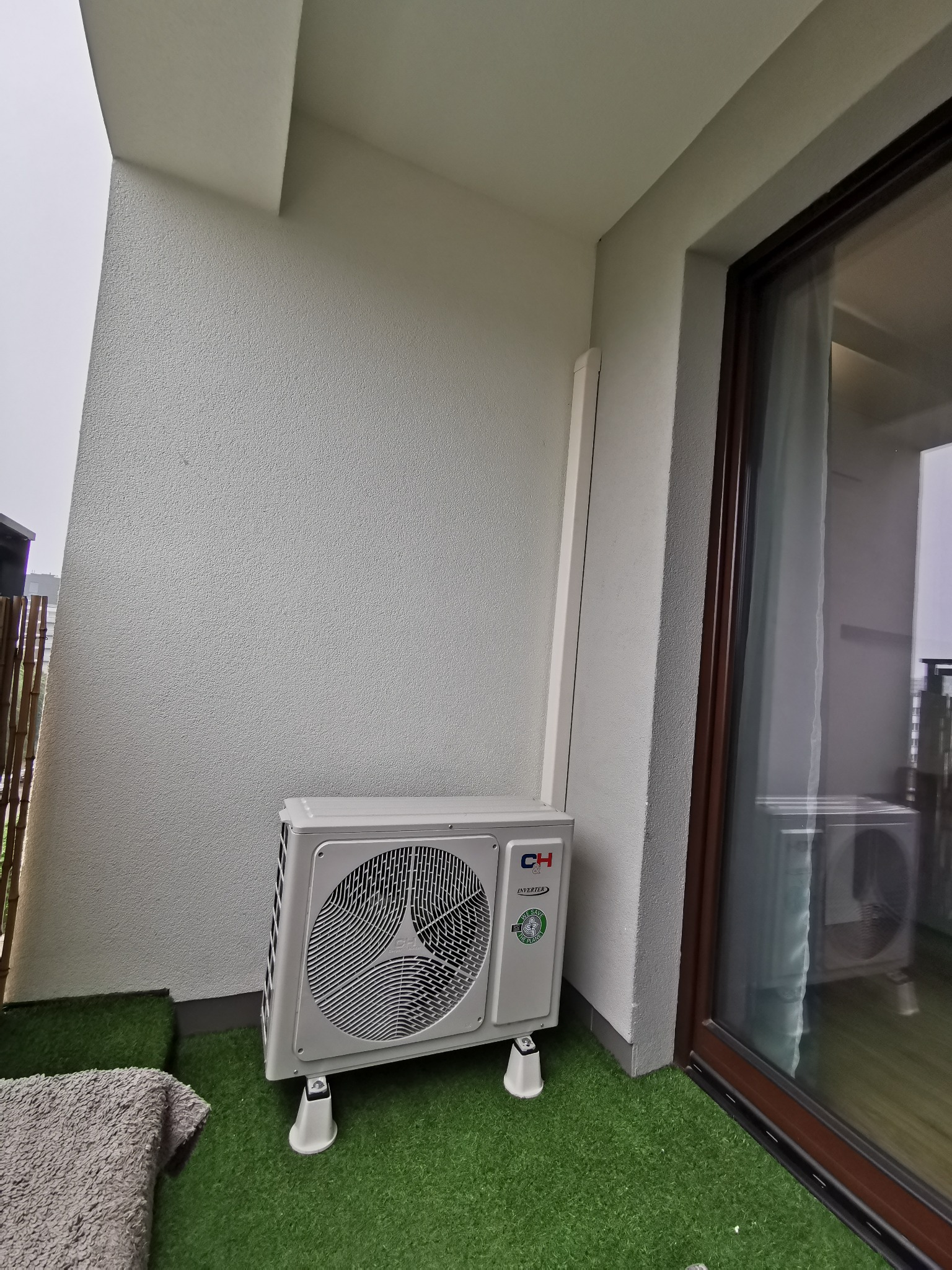 Zewnętrzna jednostka klimatyzacji CH Inverter zainstalowana na balkonie z sztuczną trawą, widoczna z bliska, z fragmentem szarego ręcznika w lewym dolnym rogu.