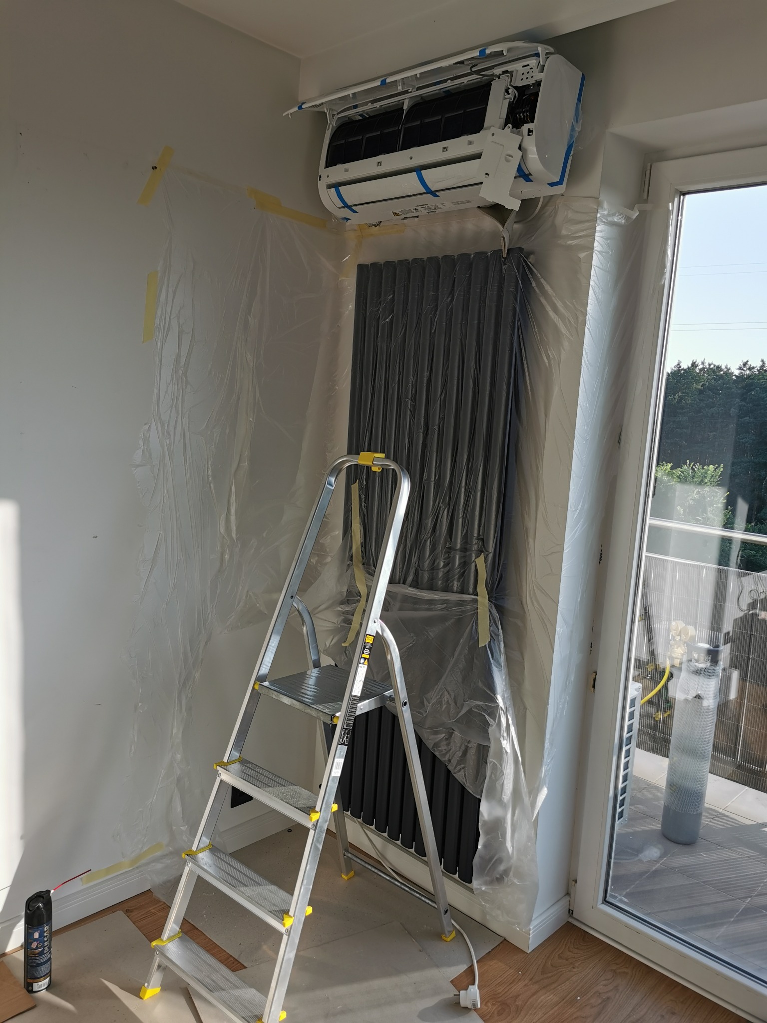 Montaż klimatyzatora w domu: widoczny nowy klimatyzator na ścianie, zabezpieczony folią grzejnik, drabina i butelka środka czyszczącego przy ścianie, przygotowania do instalacji.